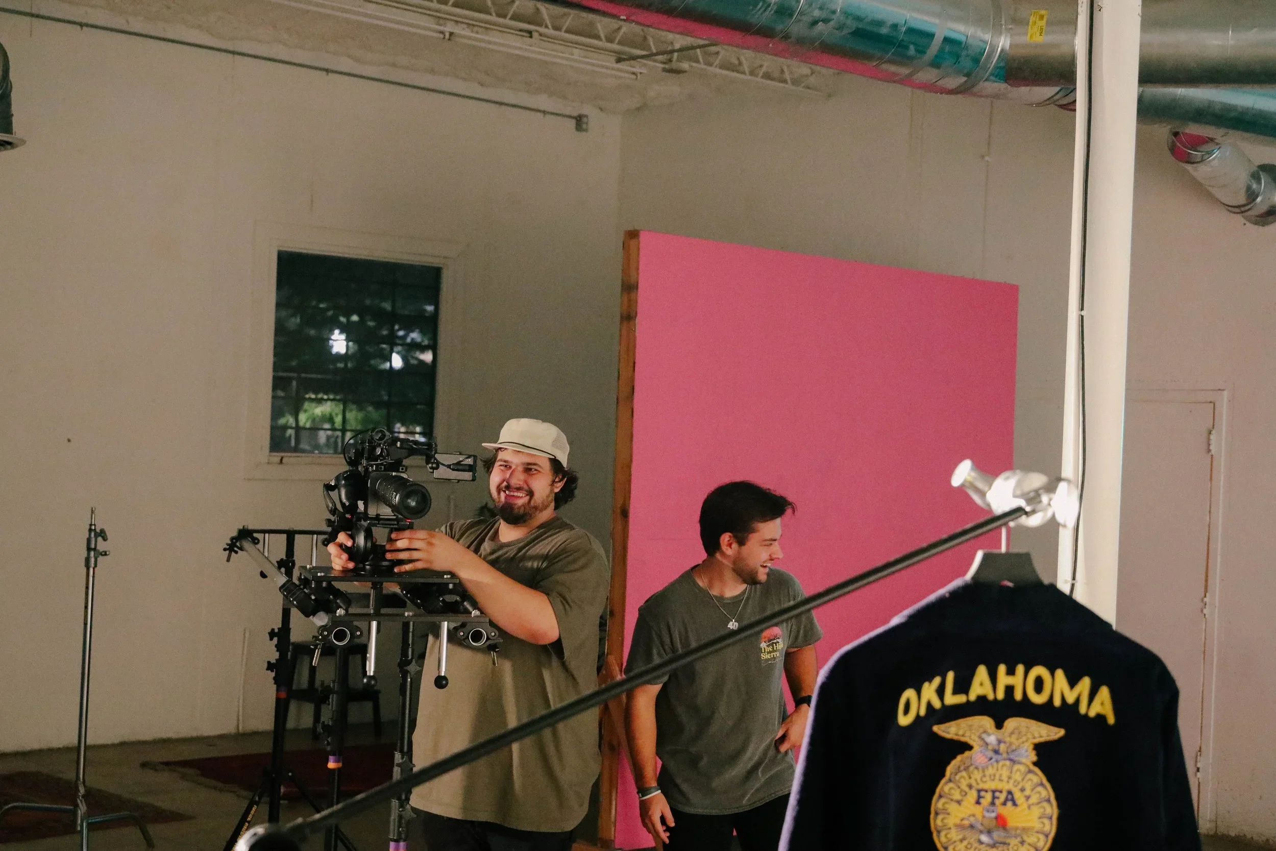 Two young men working on a film shoot, one operating a camera mounted on a tripod, and the other smiling and standing next to a pink backdrop inside a studio with industrial ceiling and a window.