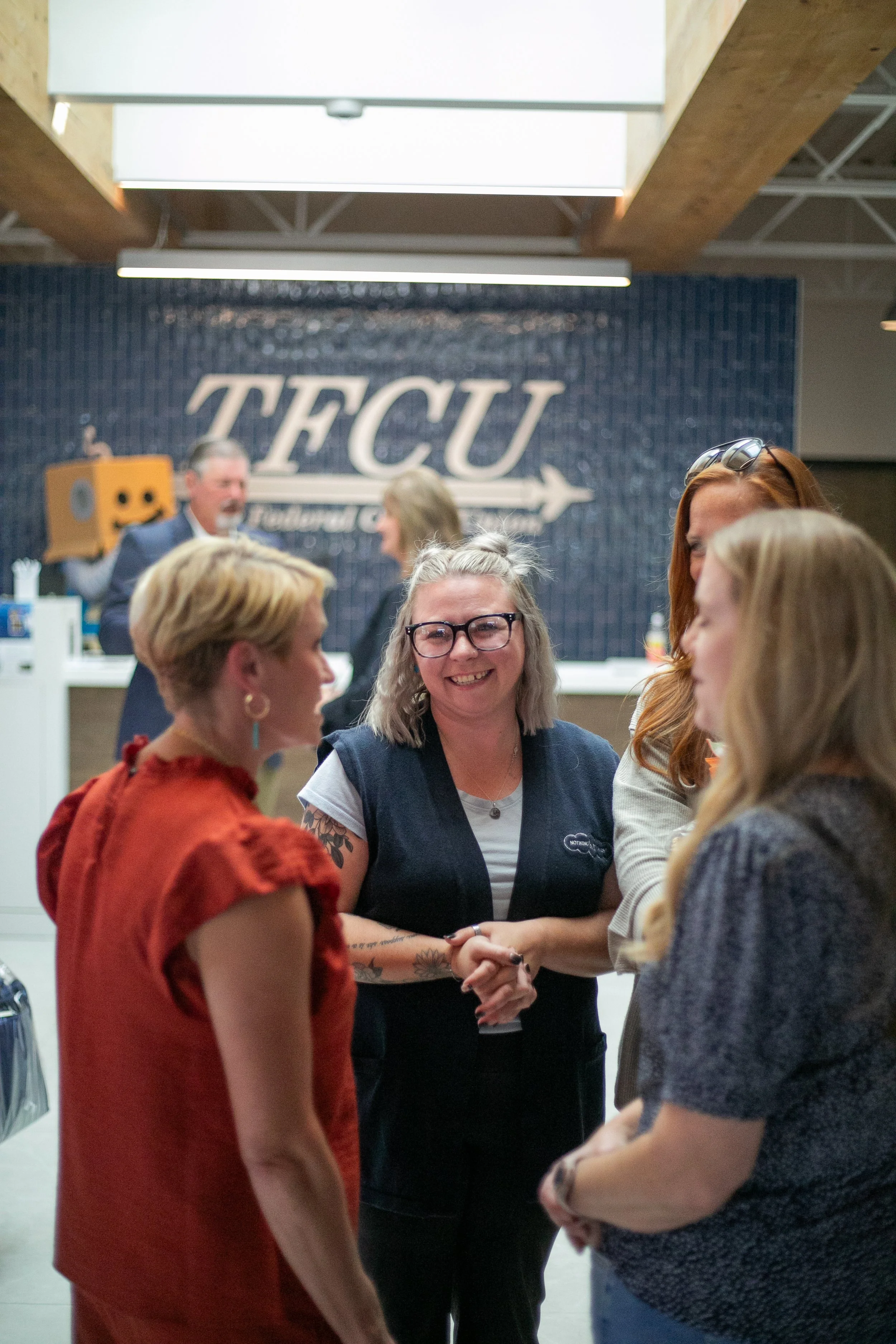 Group of women shaking hands and talking at TFCU event in front of a blue wall with the TFCU logo.