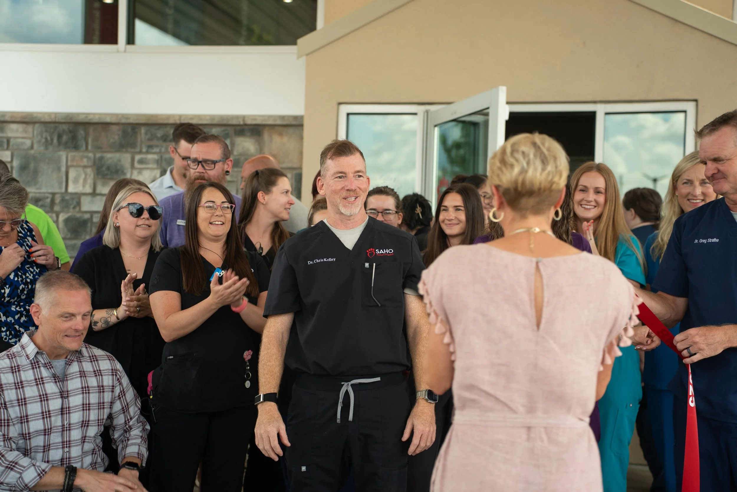 Group of healthcare professionals and staff gather outdoors, with one woman in a light pink dress receiving a ribbon or award as others applaud and smile.