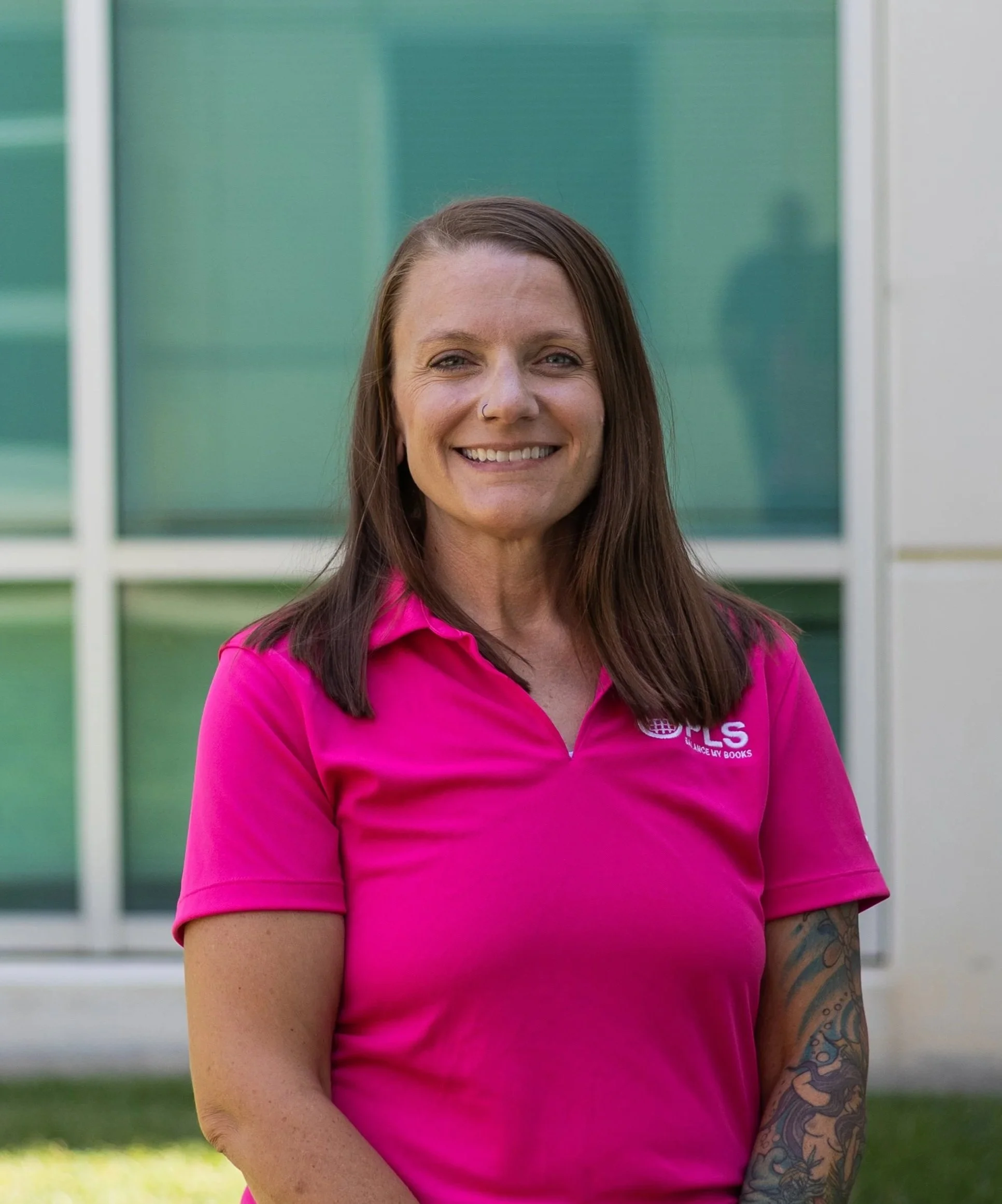 A smiling woman with shoulder-length brown hair, wearing a bright pink polo shirt with a logo on it, standing outdoors in front of a modern building with large glass windows.