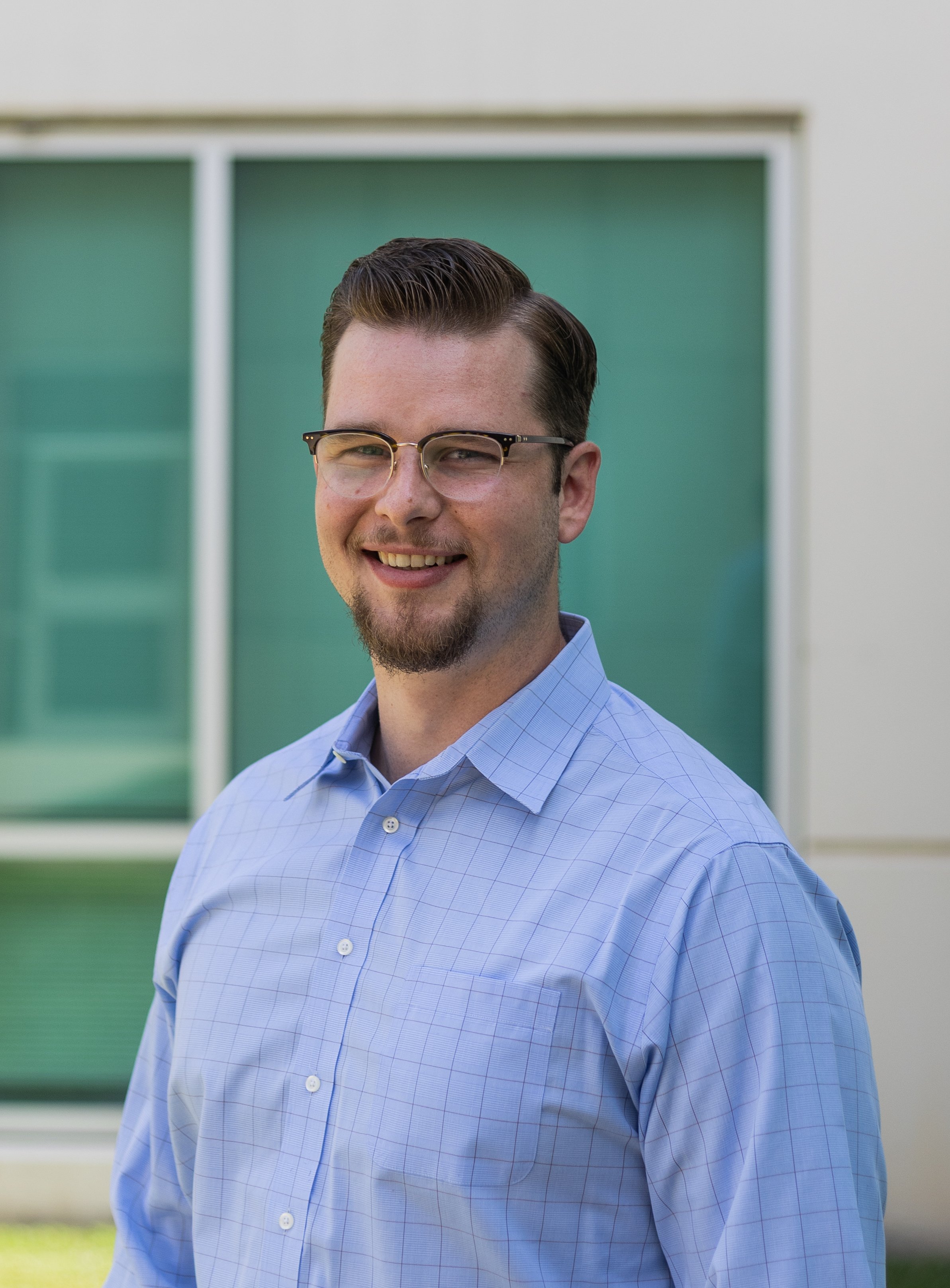 Photo of a smiling man with glasses and a beard, wearing a light blue button-up shirt, standing outdoors in front of a modern building with green-tinted windows.