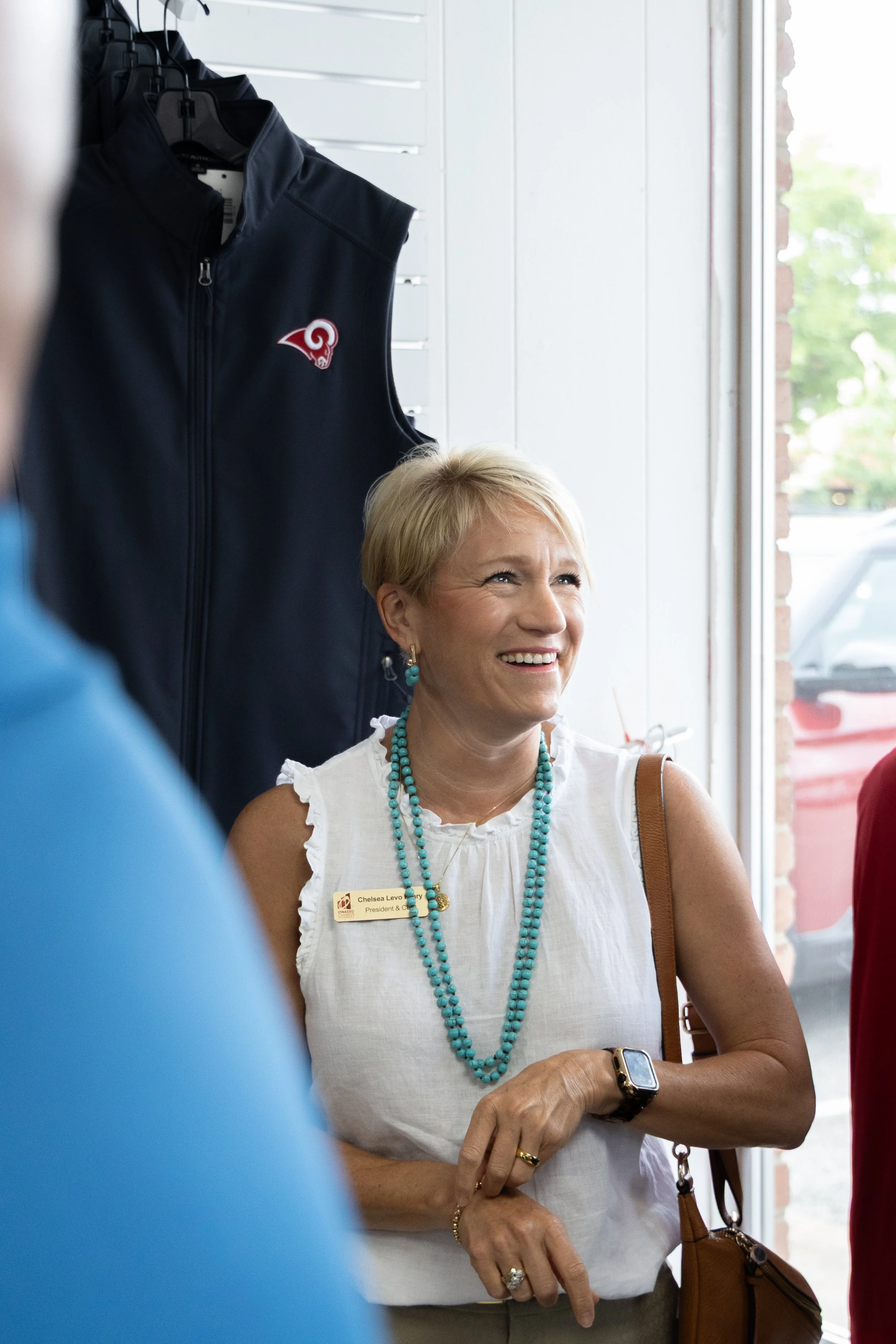 A woman with blonde hair wearing a white sleeveless top with ruffled edges, turquoise jewelry, a watch, and a name badge, smiling while standing near a large window inside a store. There is a navy blue sports vest hanging on a rack behind her.