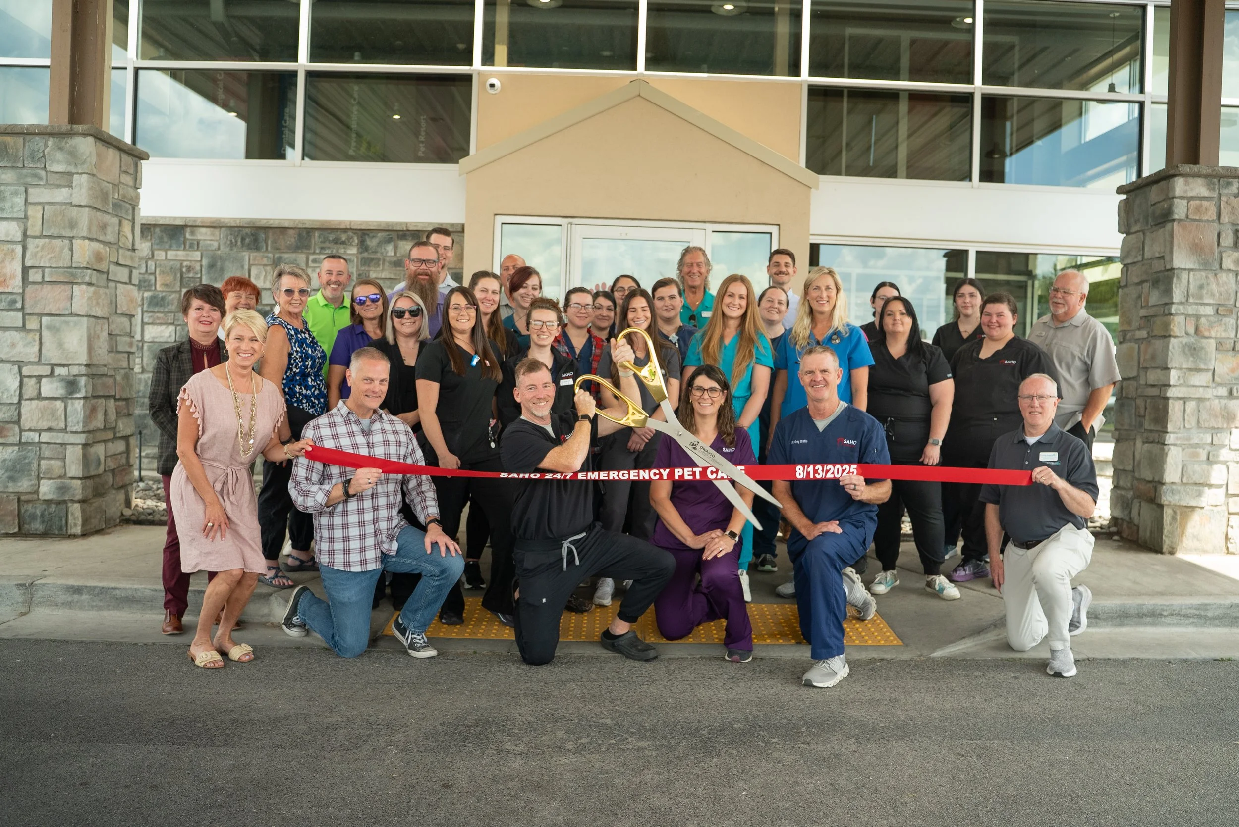 Group of staff members and community members gathered outside a building for a ribbon-cutting ceremony, with some holding large scissors and others smiling.