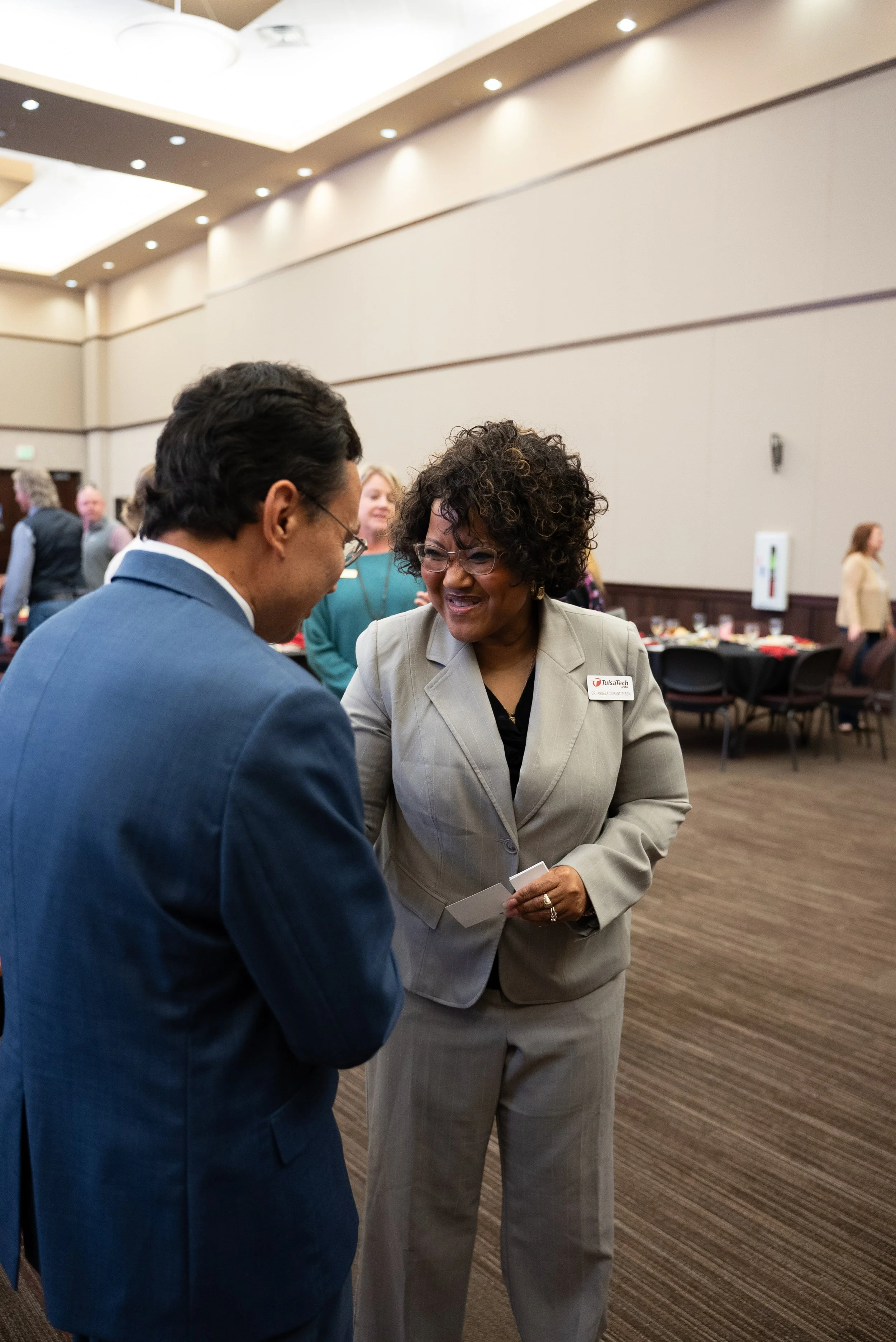 Two people, a man in a blue suit and a woman in a light gray business suit, are engaged in conversation at a professional event or conference in a large room with other attendees in the background.