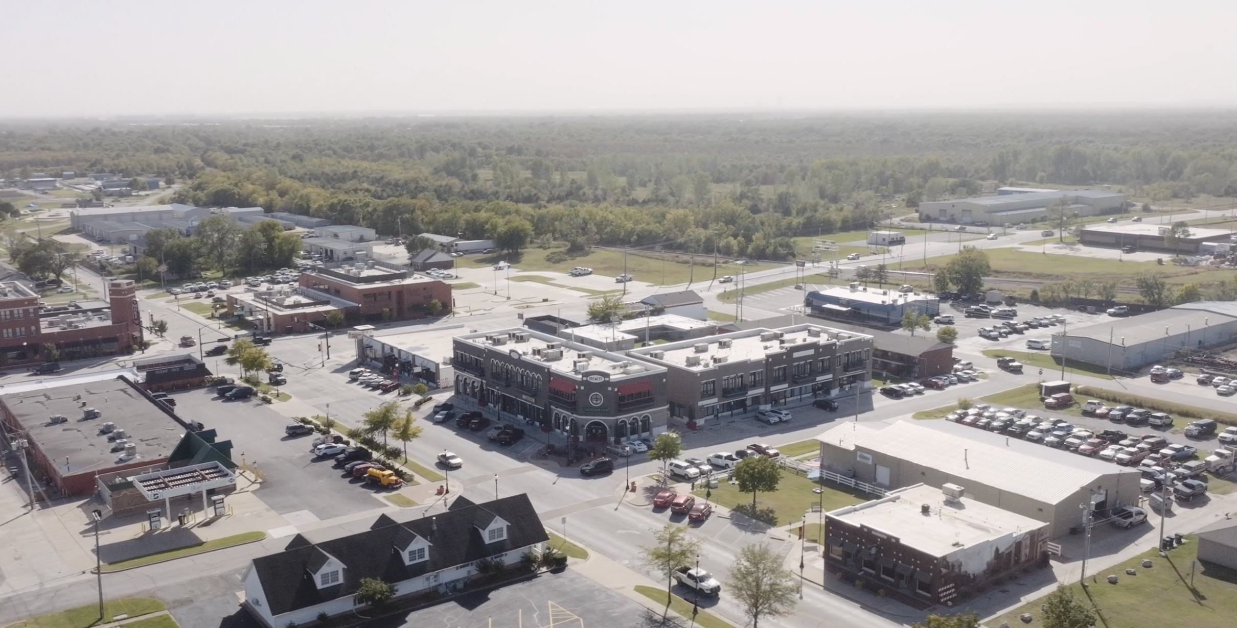 Aerial view of a small town with commercial buildings, parking lots, and surrounding green areas on a sunny day.