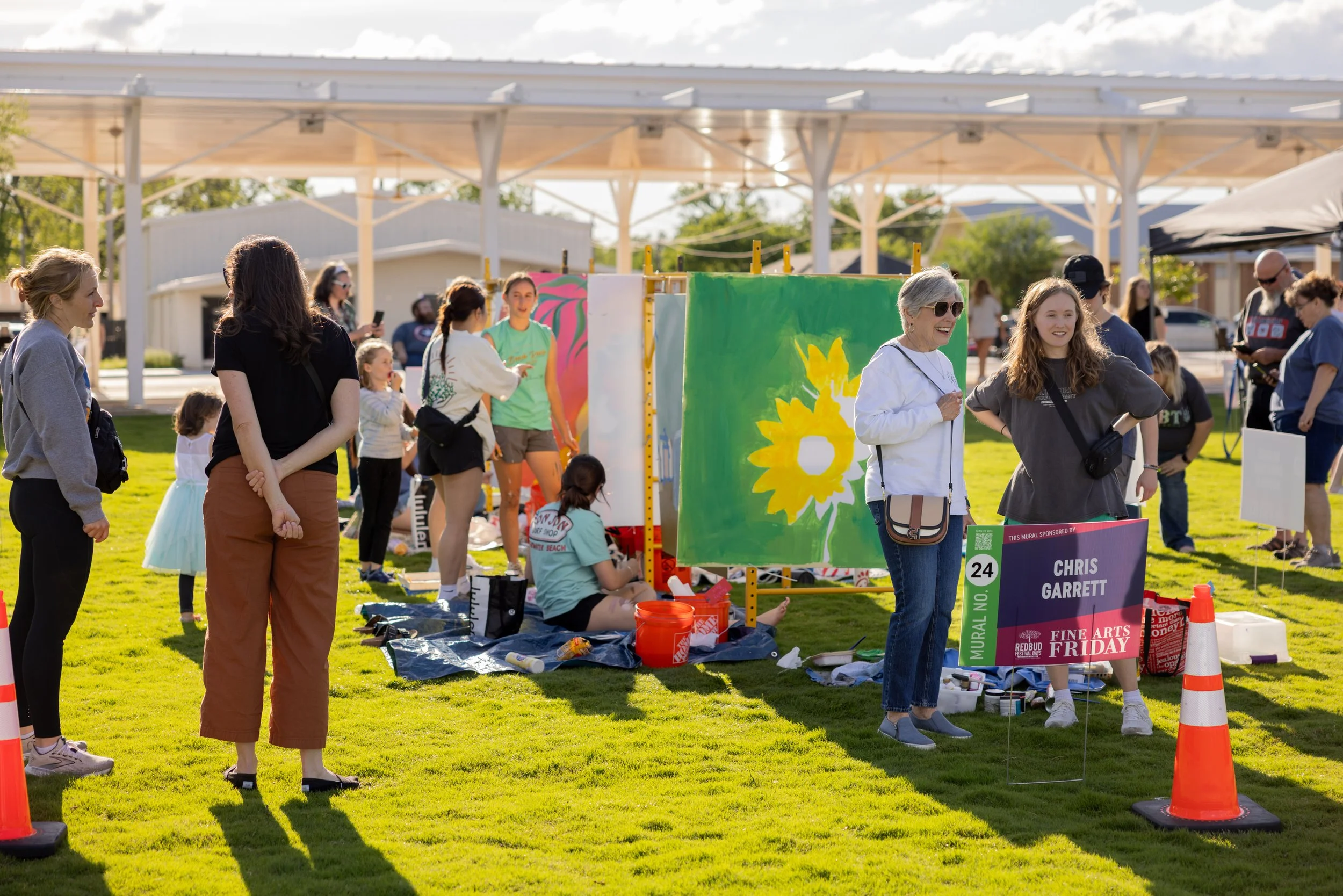 People at an outdoor art festival with displayed murals, including a vibrant floral mural, and a sign for artist Chris Garrett, in a grassy area with trees and structures in the background.