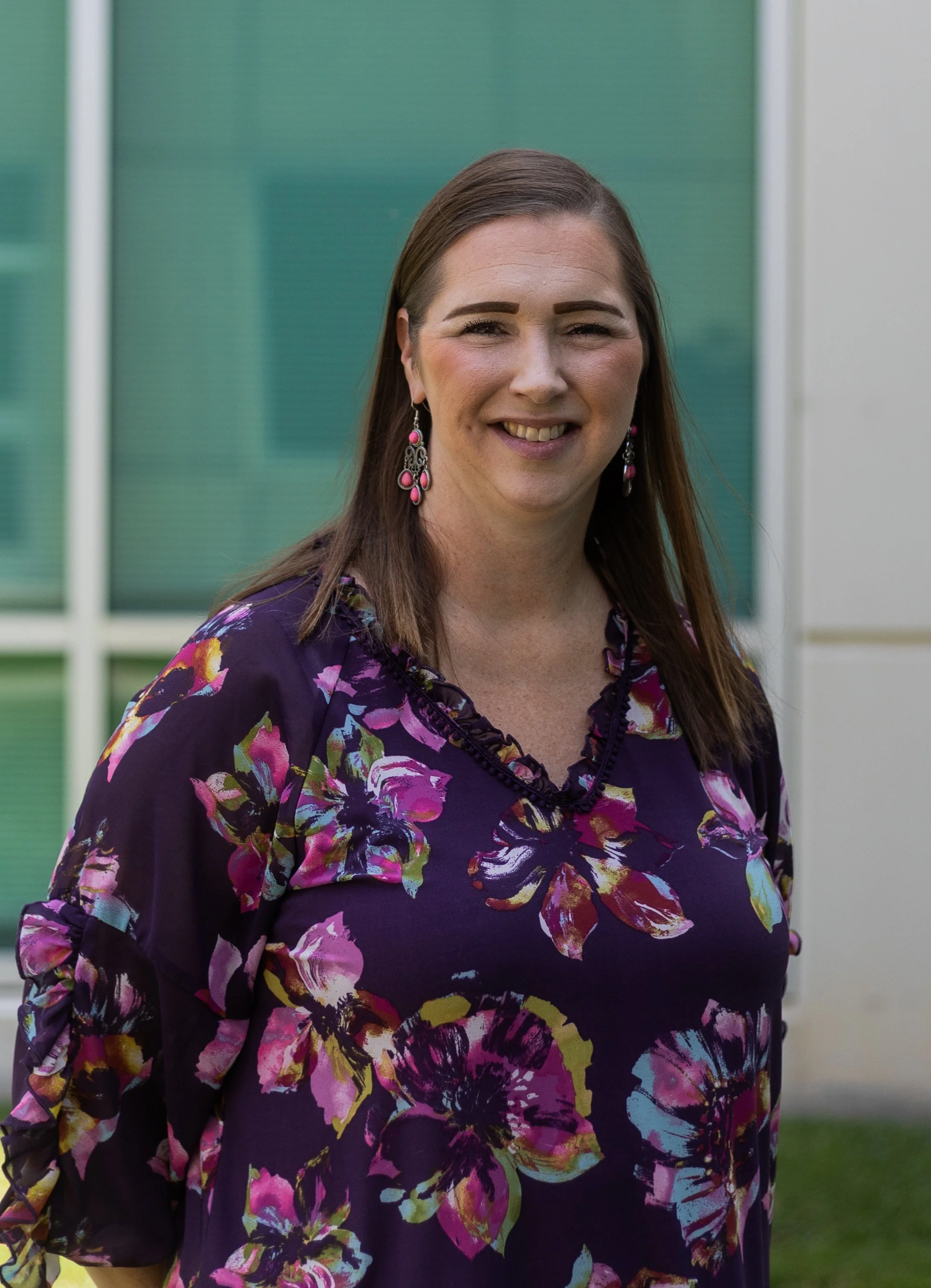 Woman with long brown hair, wearing a purple floral dress, smiling, standing outdoors with a building with green windows in the background.