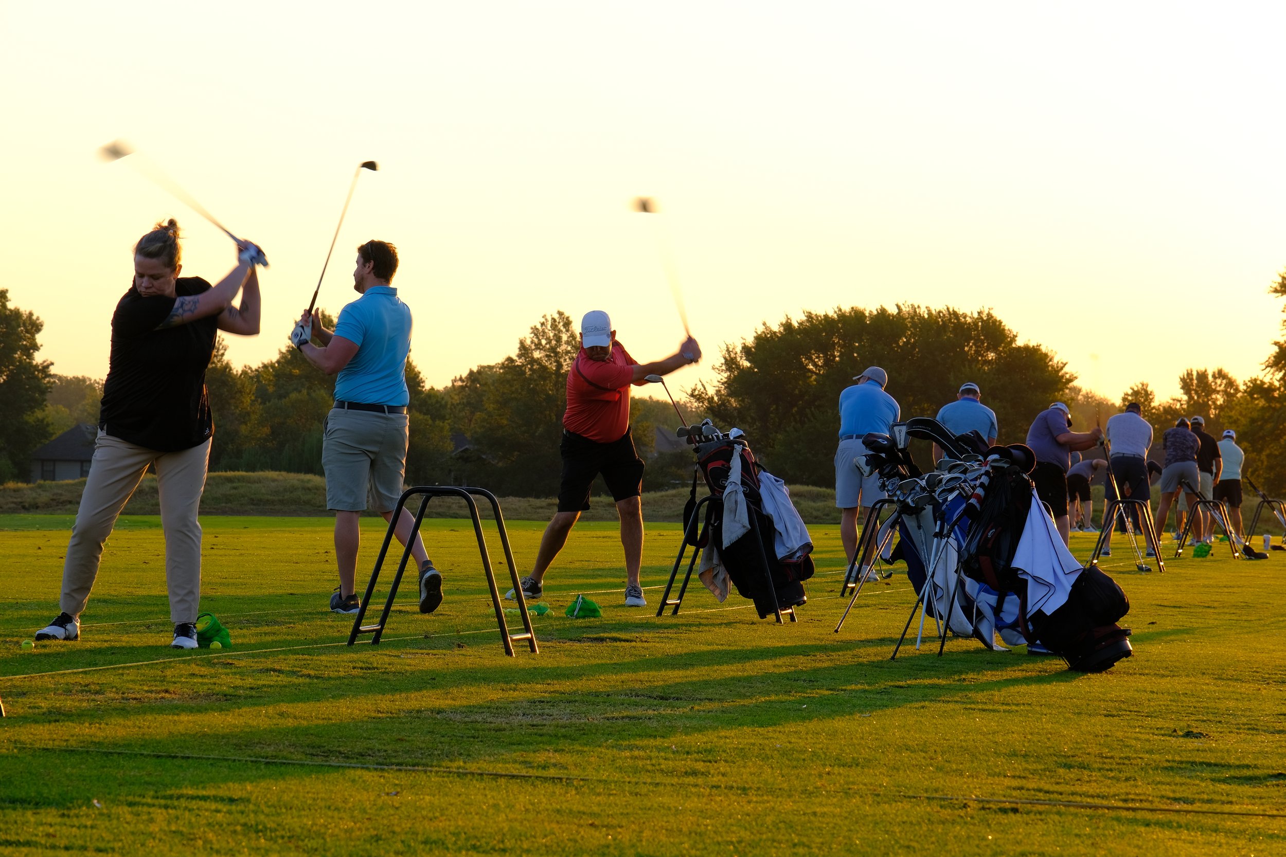 Group of golfers practicing swings on a golf driving range during sunset, with golf bags and clubs near the tee area.