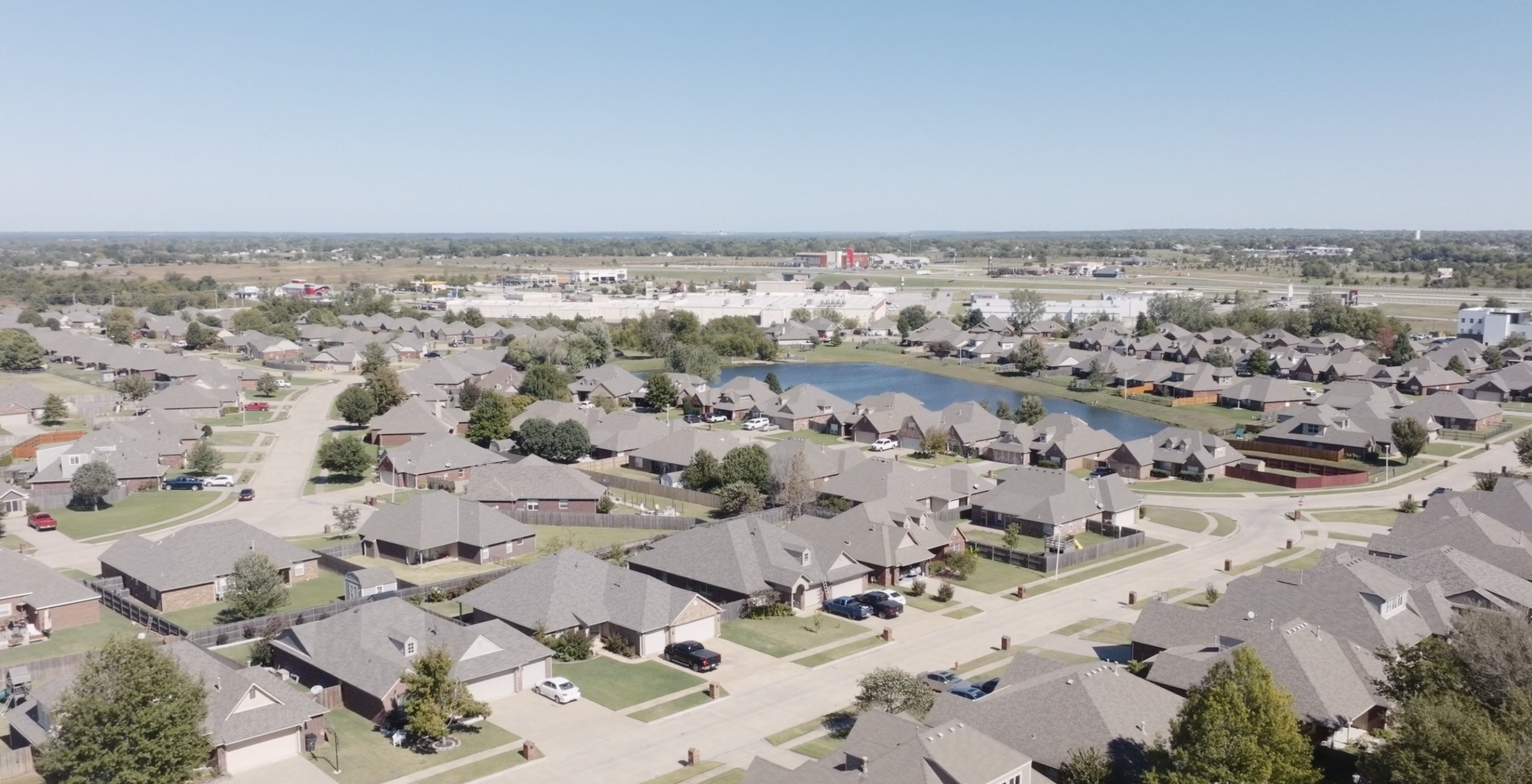 Aerial view of a suburban neighborhood with houses, trees, a pond, and streets, with commercial buildings and an industrial area in the distance.