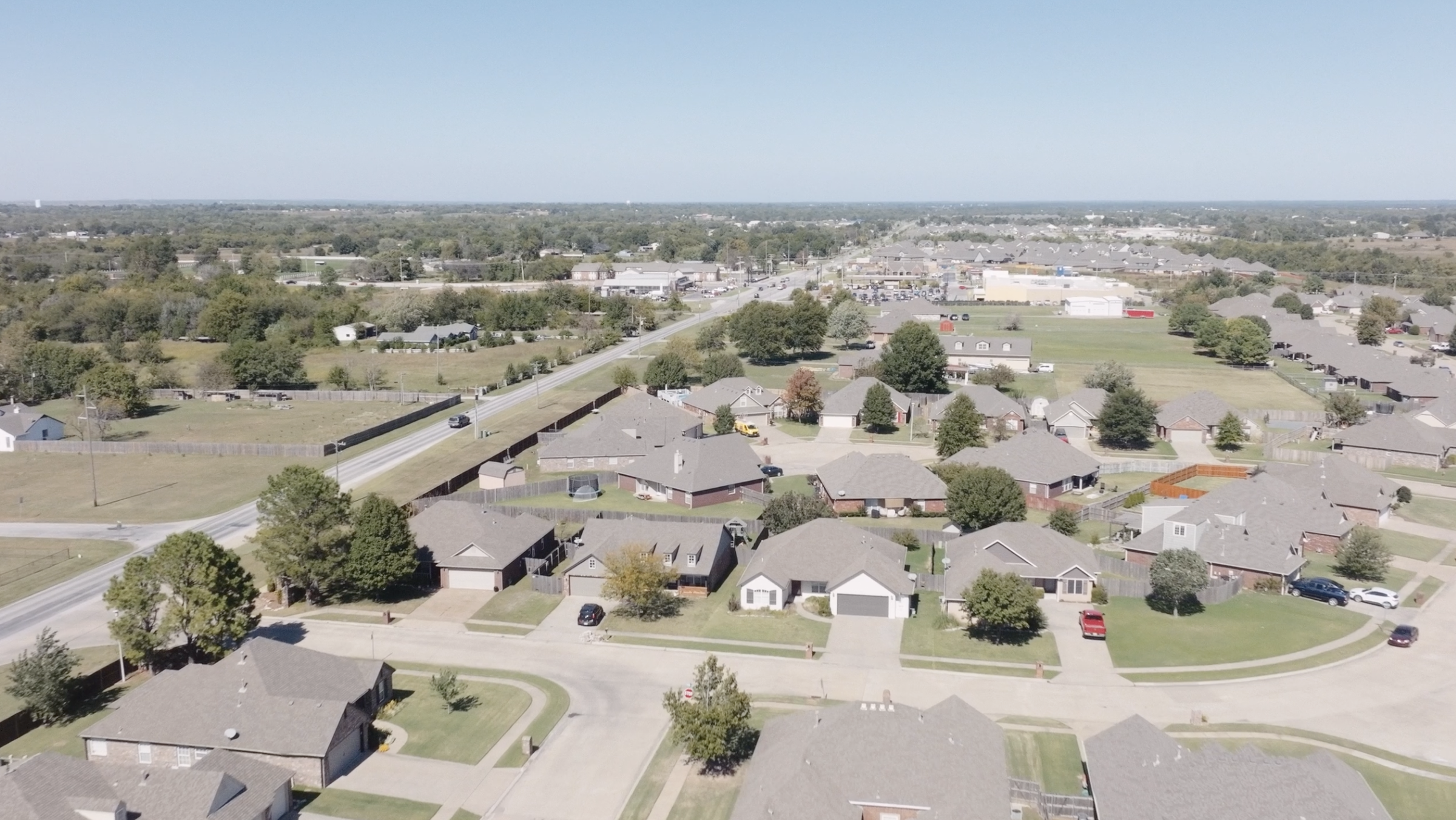 Aerial view of a suburban neighborhood with single-family homes, green lawns, trees, and a street with a car, overlooking a commercial area in the distance under a clear blue sky.