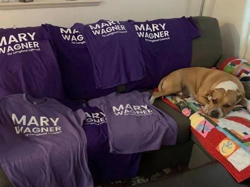 Purple campaign shirts with white text that reads 'Mary Wagner for Longford Council' draped over a gray couch, alongside a sleeping brown and white dog on a colorful quilt.