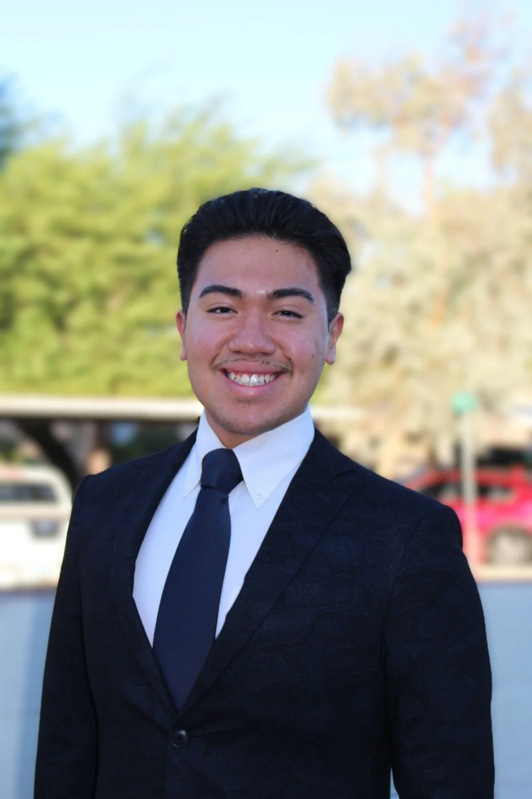 A young man in a dark suit and tie smiling outdoors with trees and parked cars in the background.