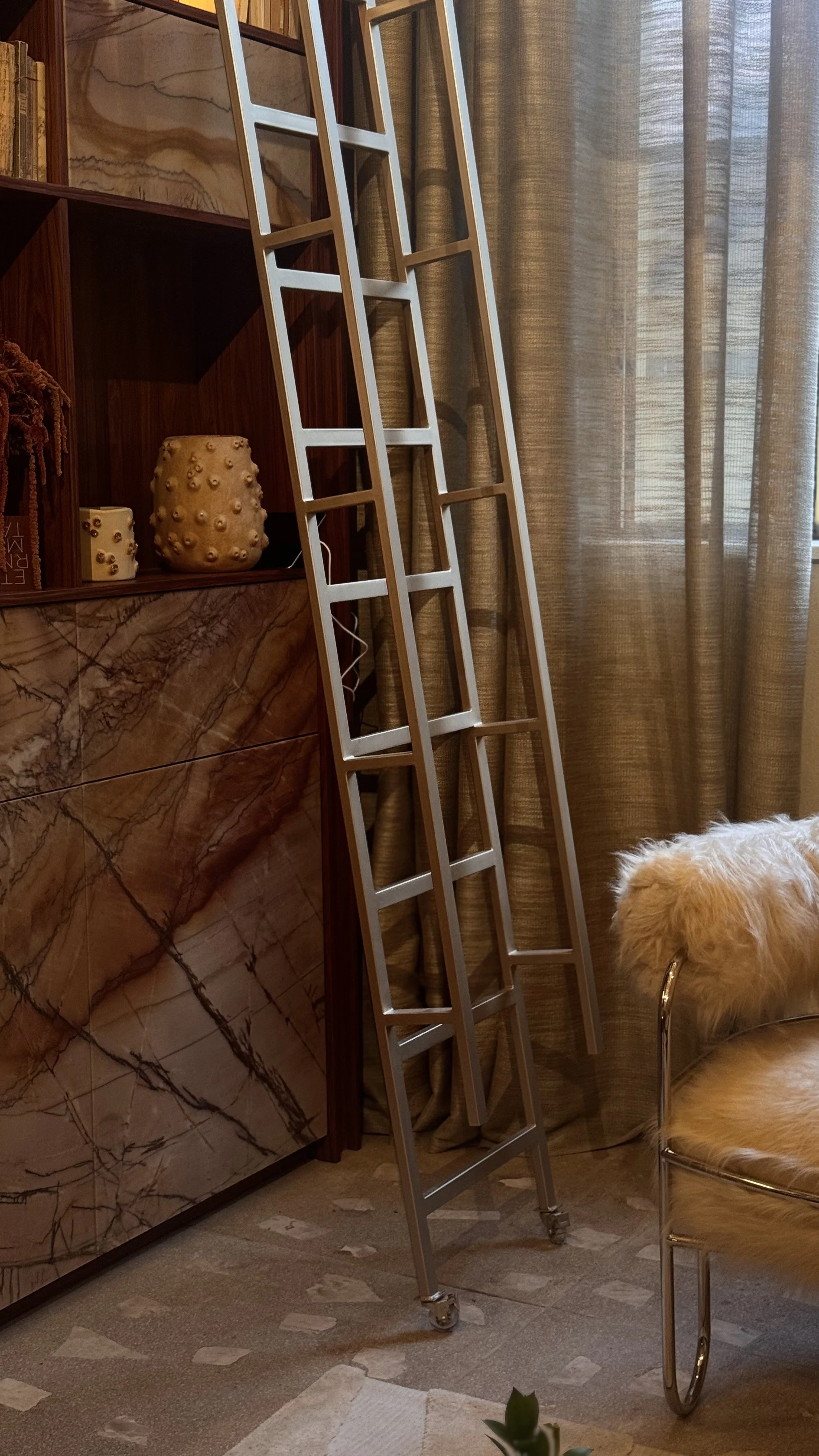 Uma escada de metal prateada apoiada na parede de um ambiente interno, perto de cortinas e de um móvel de madeira com uma superfície de mármore com veios escuros. Há uma cadeira com encosto de pelúcia ao lado da escada.