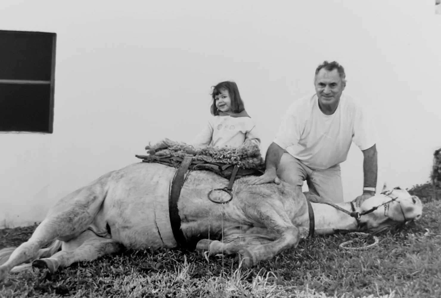 Homem e menina com um cavalo morto, na grama, em uma parede branca, em preto e branco.