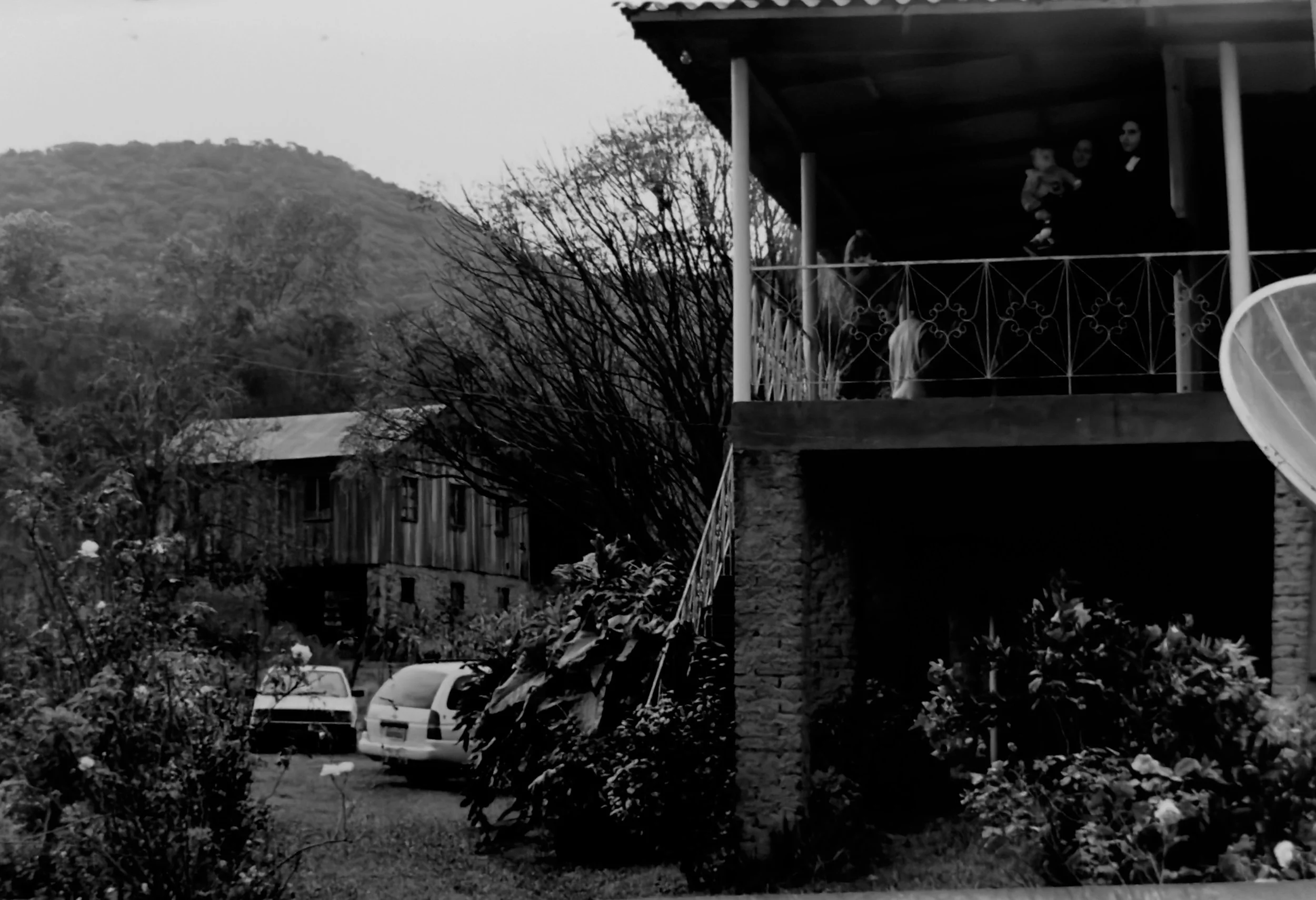 Vista de uma casa com varanda no segundo andar, onde há duas pessoas, uma criança e uma mulher, observando para baixo. No jardim, há várias plantas e uma estrada com três carros estacionados. No fundo, há uma colina com árvores.