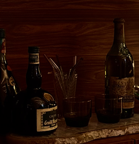 Garrafas de bebida alcoólica, copos com bebida escura e um recipiente com palitos de vidro sobre uma bancada de madeira.