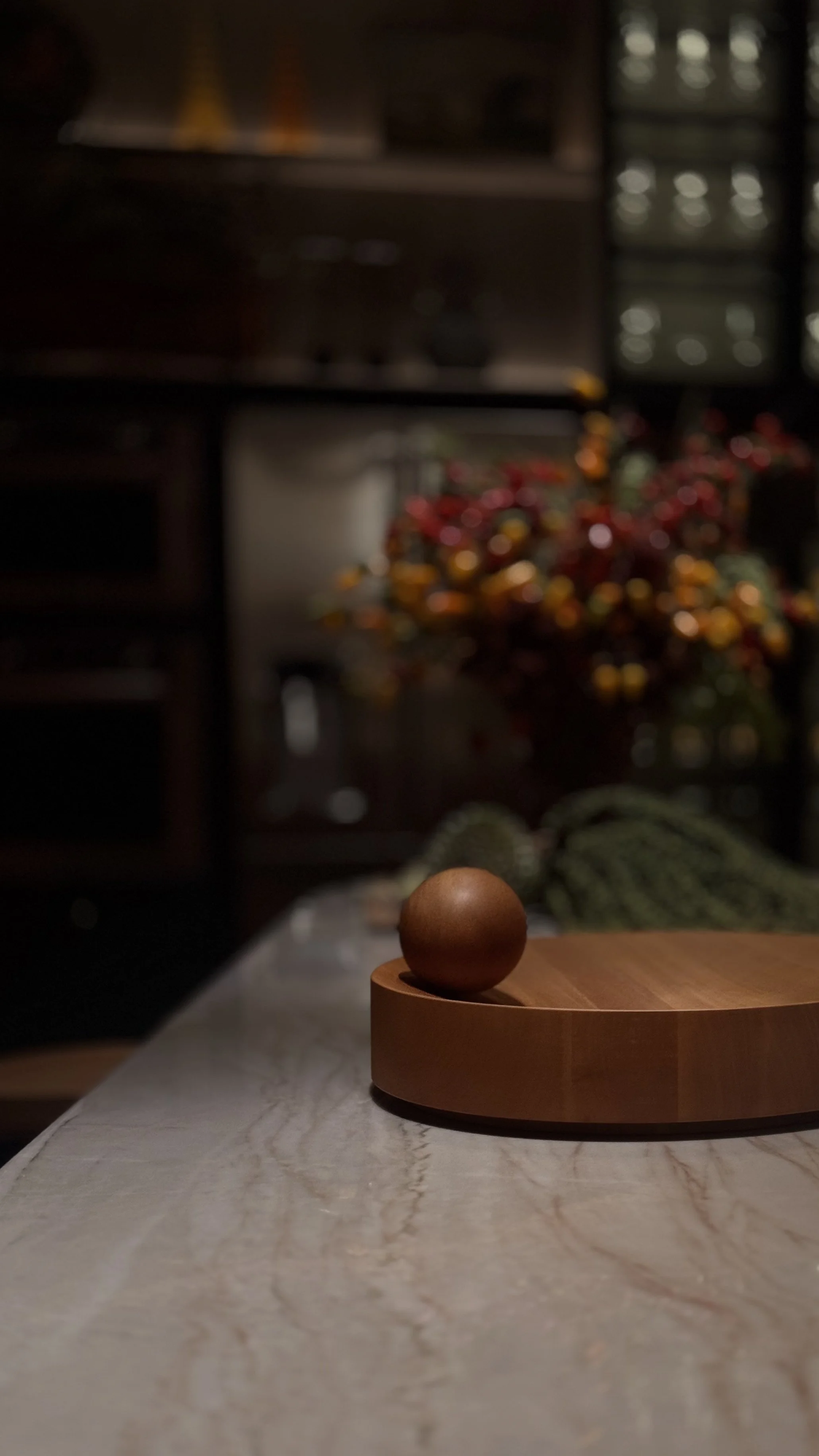 Uma mesa de mármore com uma tábua de madeira redonda e uma bola de cera ou argila de cor marrom. Ao fundo, uma decoração de Natal com bolas vermelhas, douradas e verdes, e uma janela com vidro fumê.