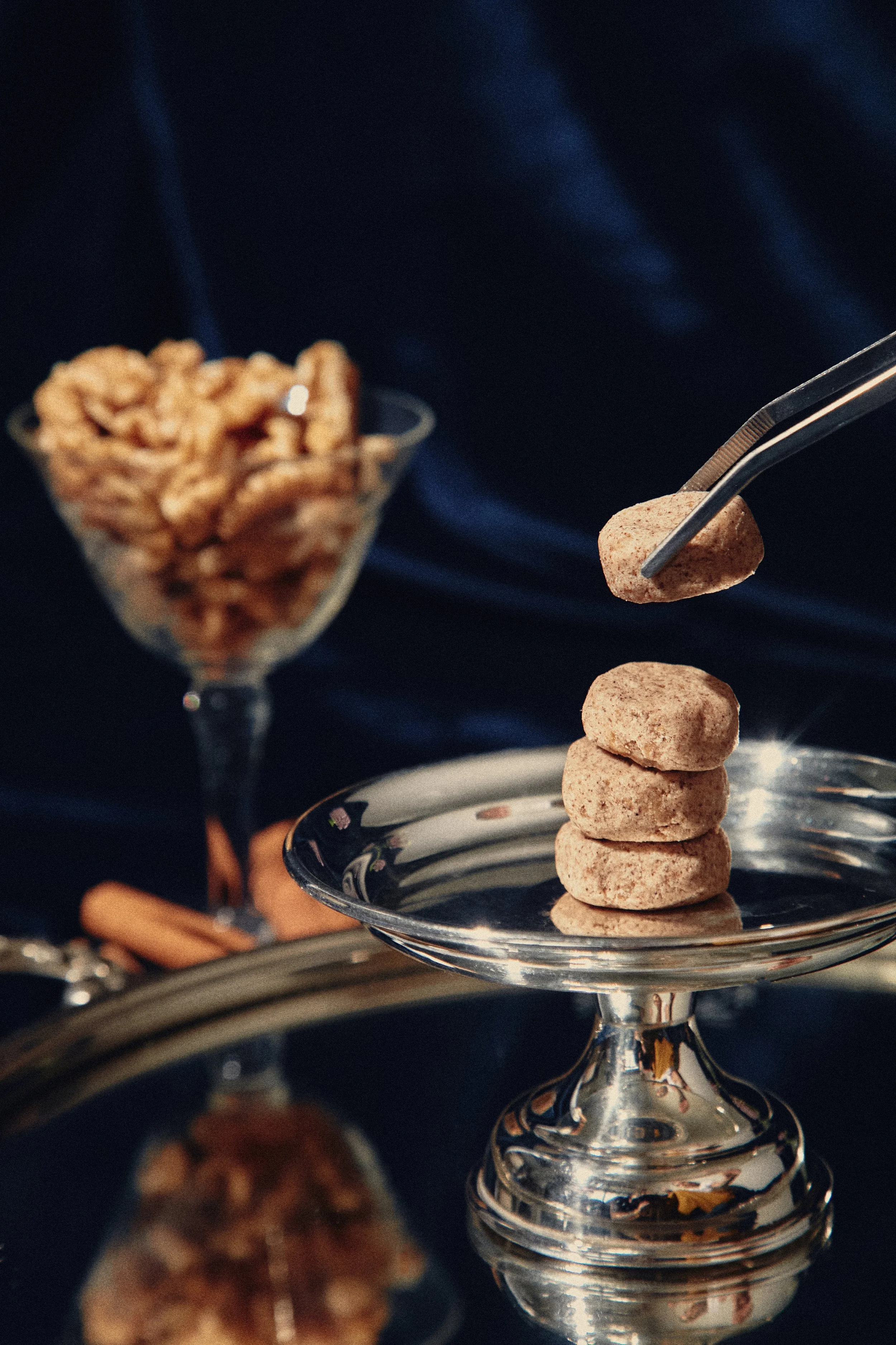 Três biscoitos empilhados em um prato de prata, com uma pinça segurando um biscoito acima. Ao fundo, um copo de taça mostra um recipiente de nozes ou castanhas.