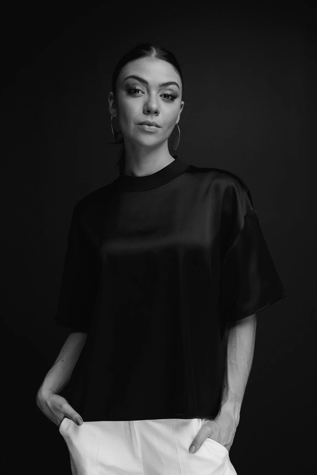 Retrato em preto e branco de uma mulher com cabelo escuro, usando uma blusa preta de tecido brilhante, brincos grandes circulares e calças brancas, posando com as mãos nos bolsos contra um fundo escuro.