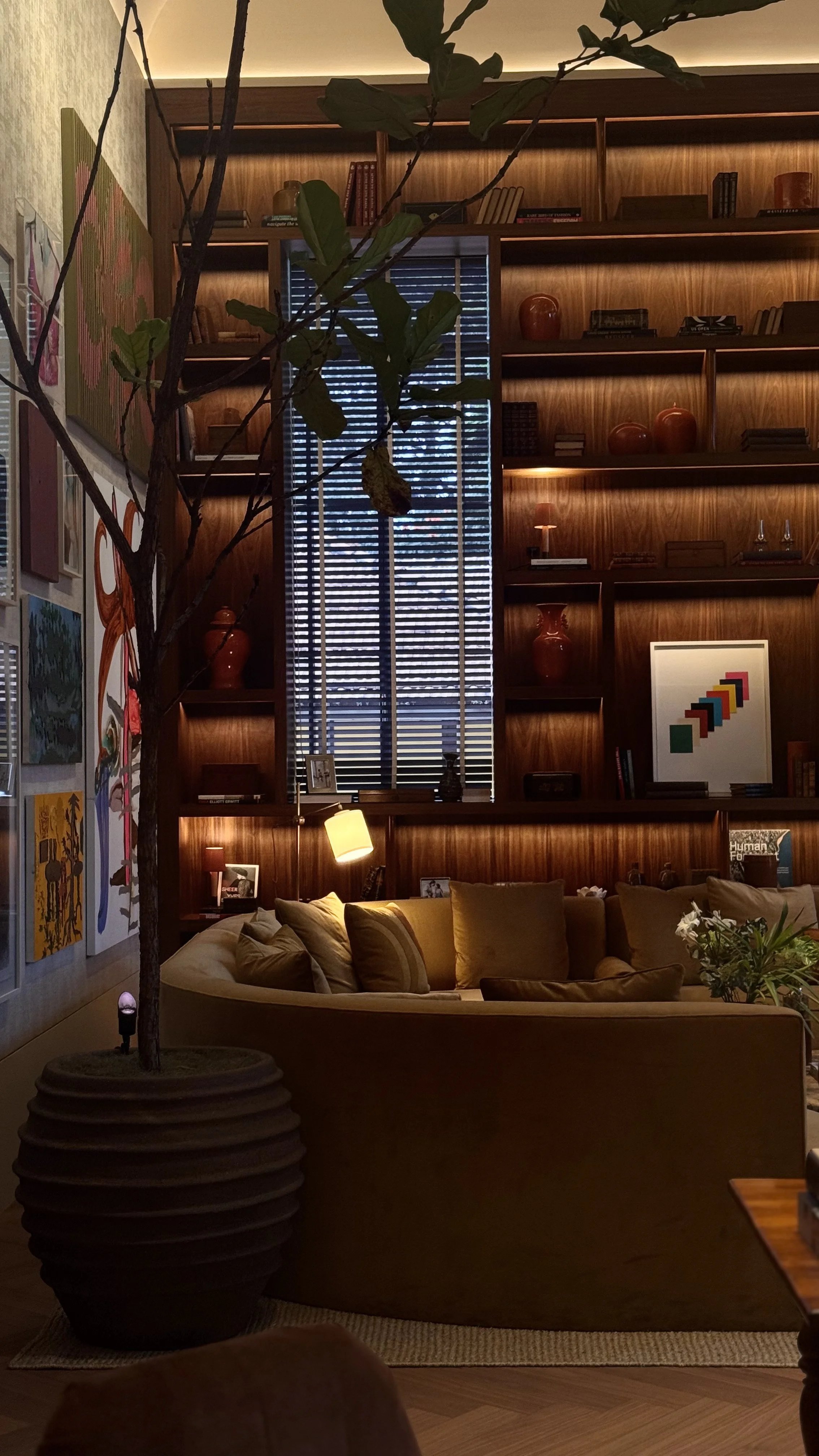 Interior de uma sala aconchegante com uma grande planta em vaso escuro, uma parede de prateleiras de madeira com livros, vasos e objetos decorativos, uma janela com persianas, um sofá de canto com almofadas, e iluminação suave.