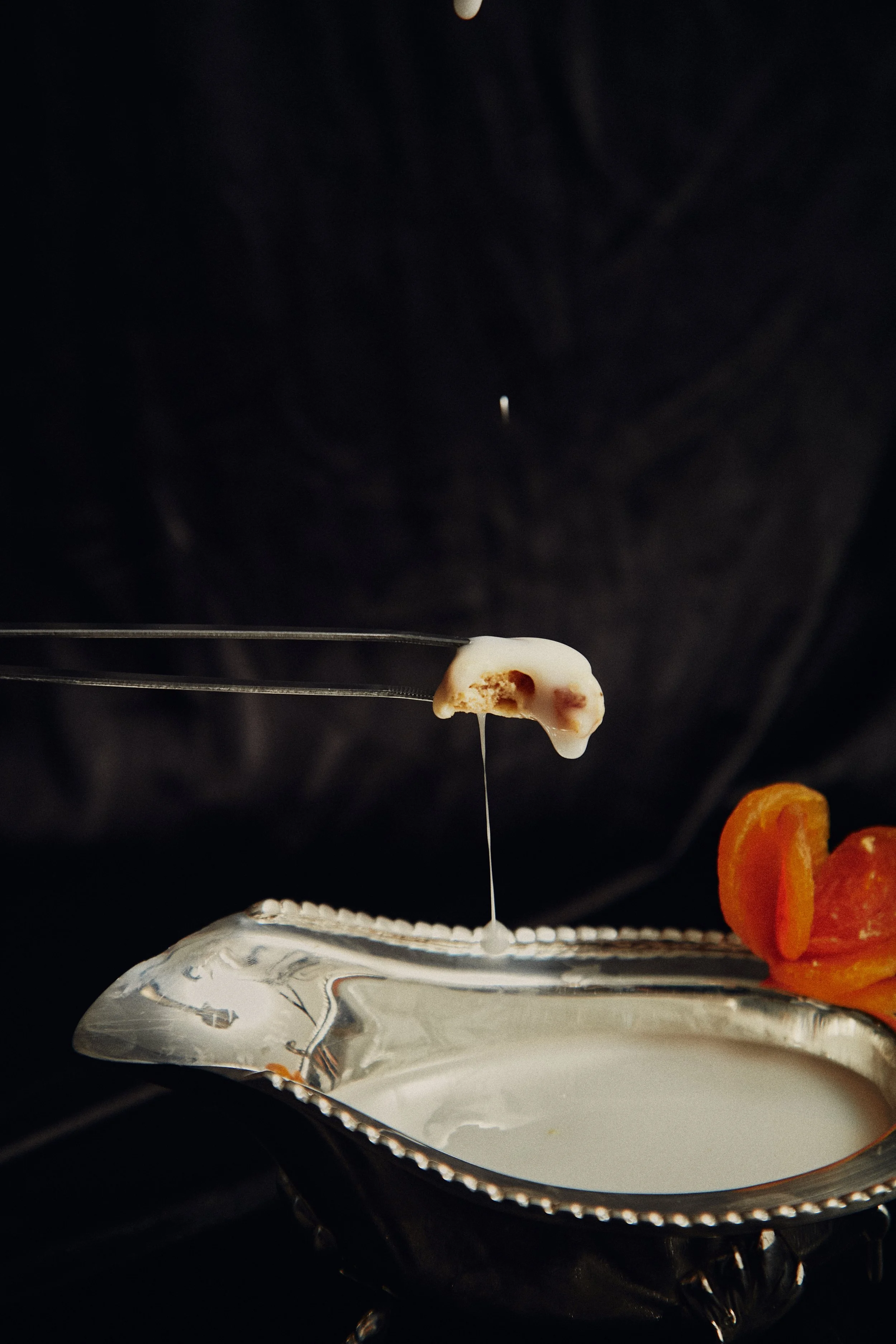 Queijo derretido em uma colher sendo mergulhado em uma tigela com molho branco, ao lado de damascos secos.