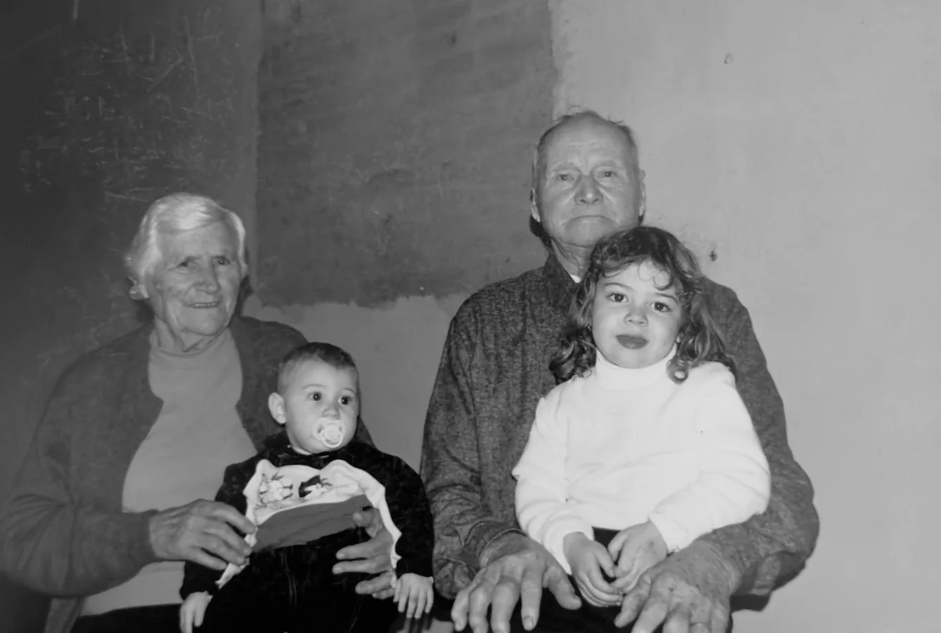 Imagem em preto e branco de três gerações: uma avó, um avô e dois jovens, possivelmente netos, sentados juntos por trás de uma parede simples.