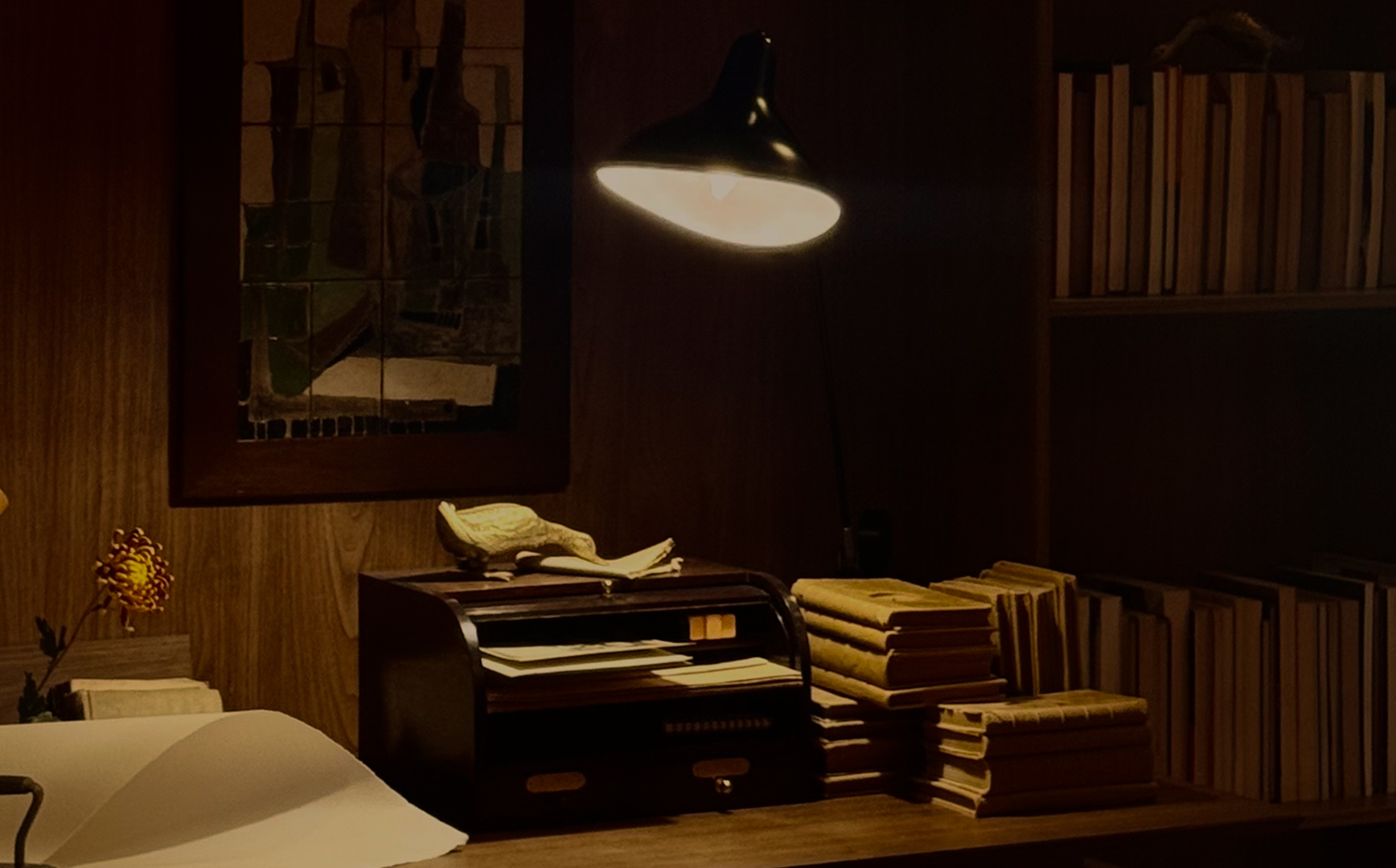 Estante de madeira com pilhas de livros, uma luminária acesa, uma flor no canto, uma tela na parede com uma pintura e objetos decorativos na mesa ao lado.