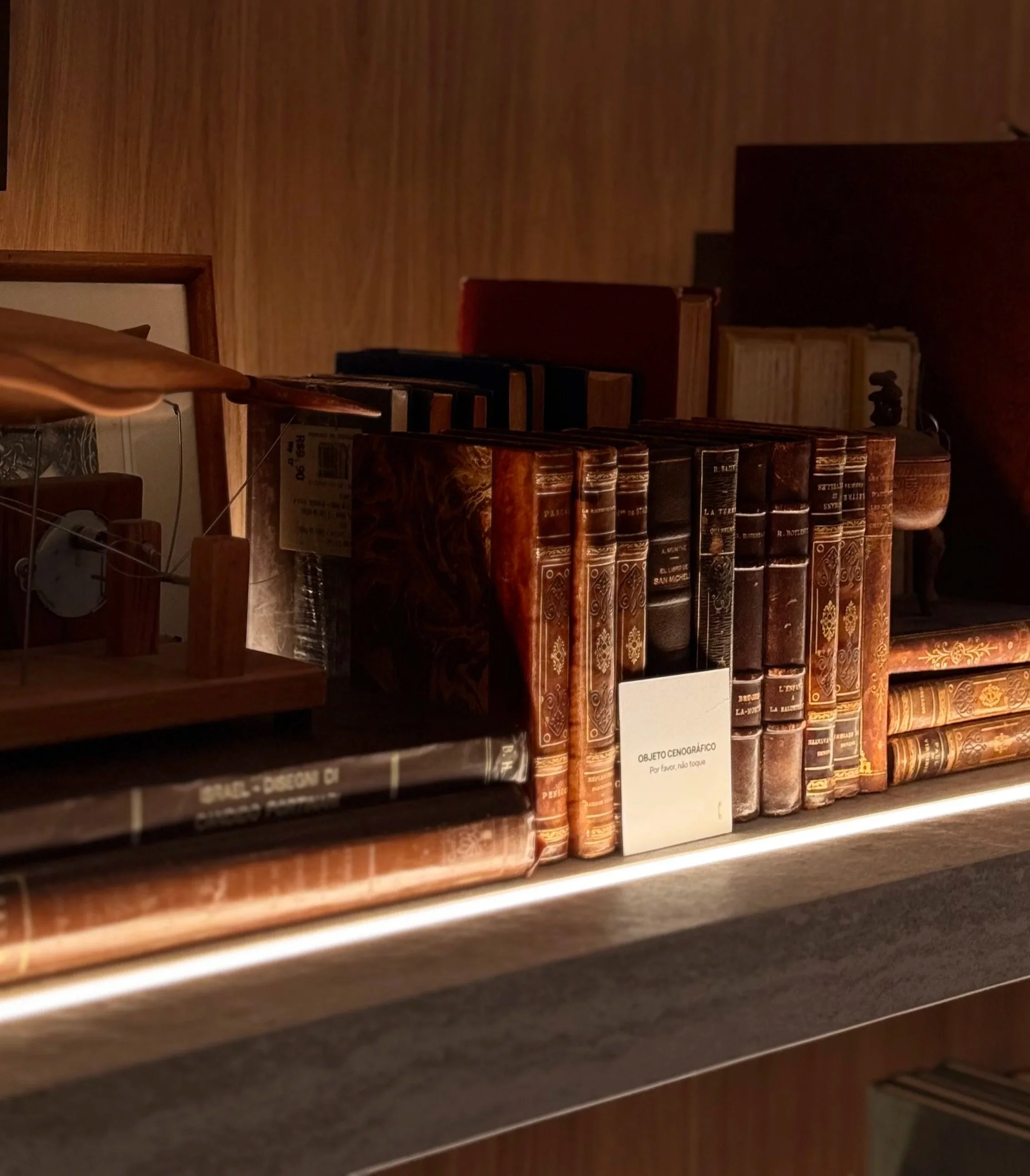 Estante com livros antigos de capa dura e cor marrom, com uma placa identificadora na frente que diz 'Objeto Cenográfico'.