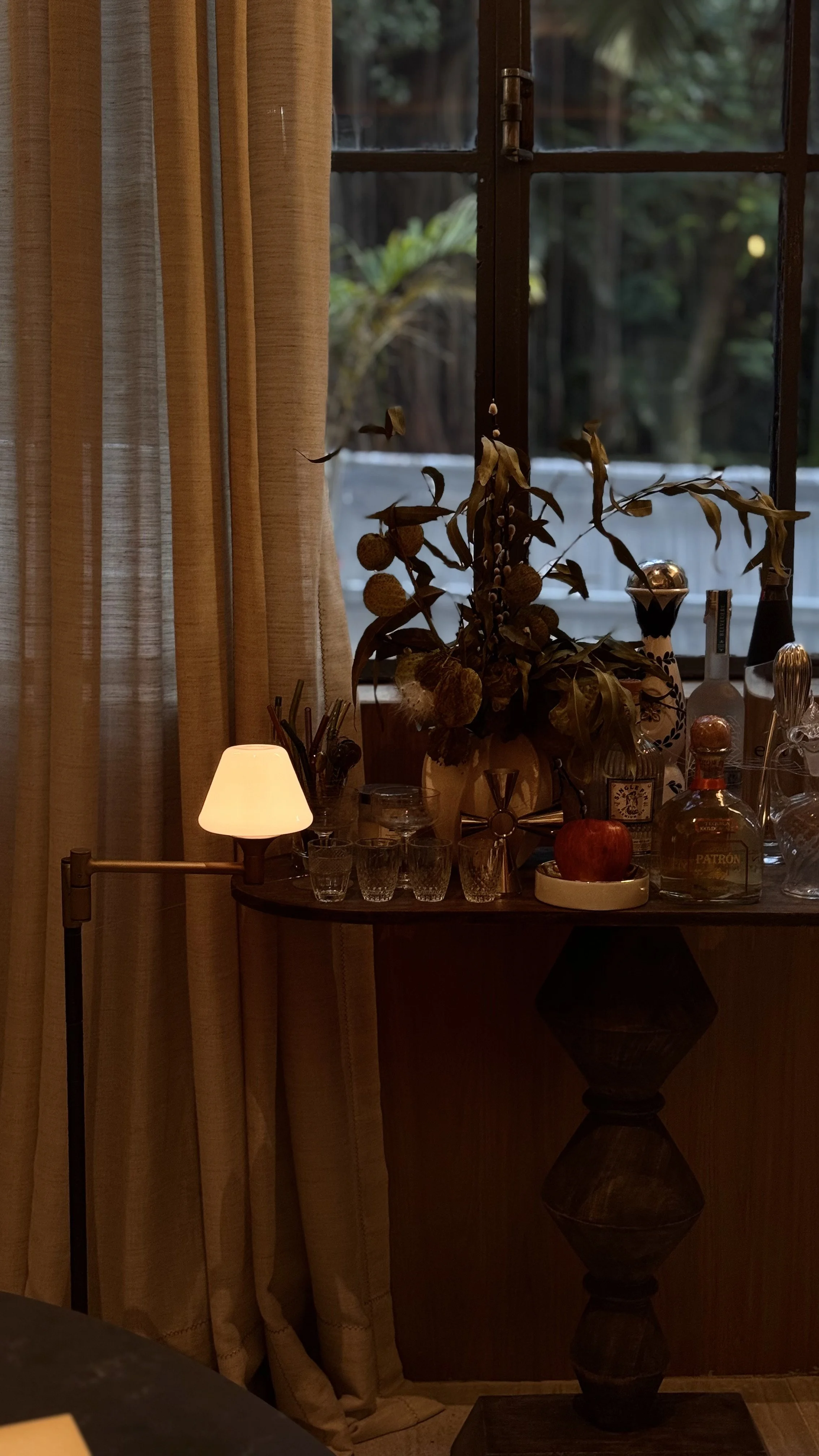 Vista de uma mesa de bar com várias garrafas, copos e uma planta seca decorativa ao lado de uma janela com cortinas em um ambiente interno, com luz suave