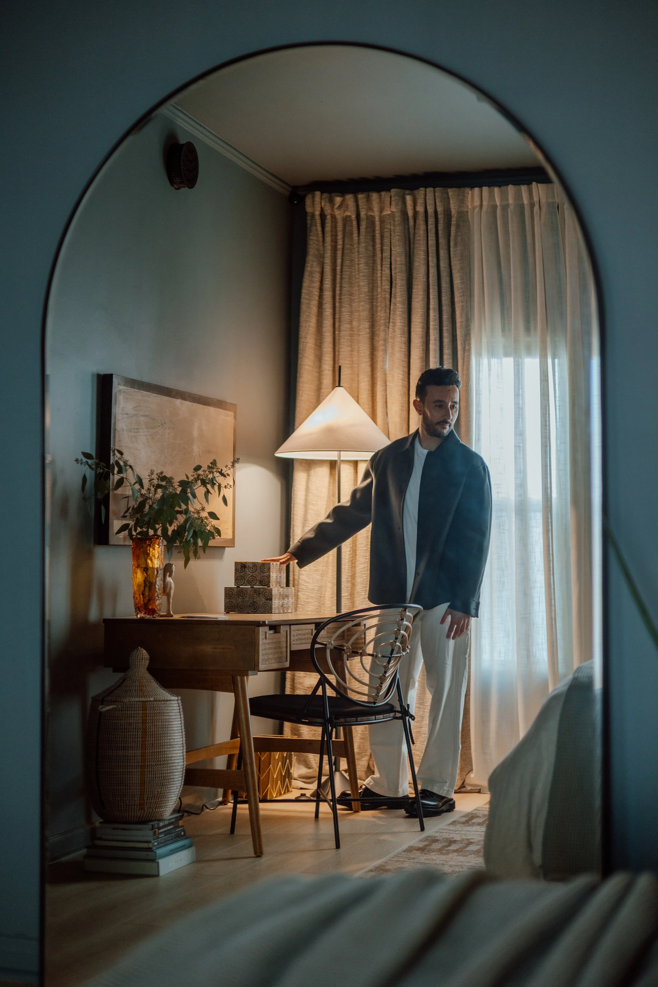 A man standing in a cozy room, viewed through an arched mirror. He is near a window with curtains, beside a desk with decorative items and a potted plant. The room is warmly lit with a floor lamp.