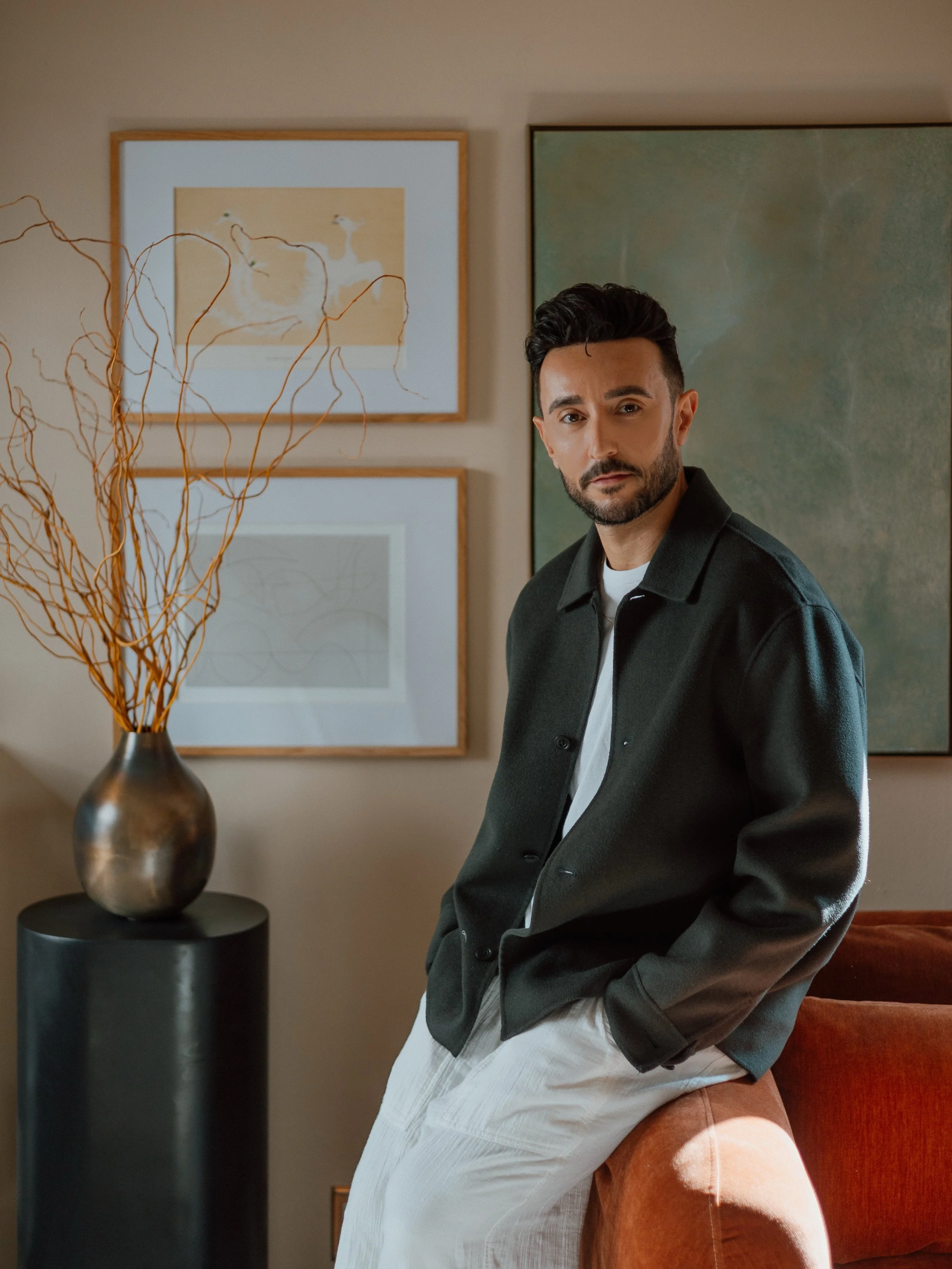 A man with dark hair and a beard leaning against a beige armchair, wearing a dark jacket and light pants, in a modern living room with framed art on the wall and a decorative vase on a stand.