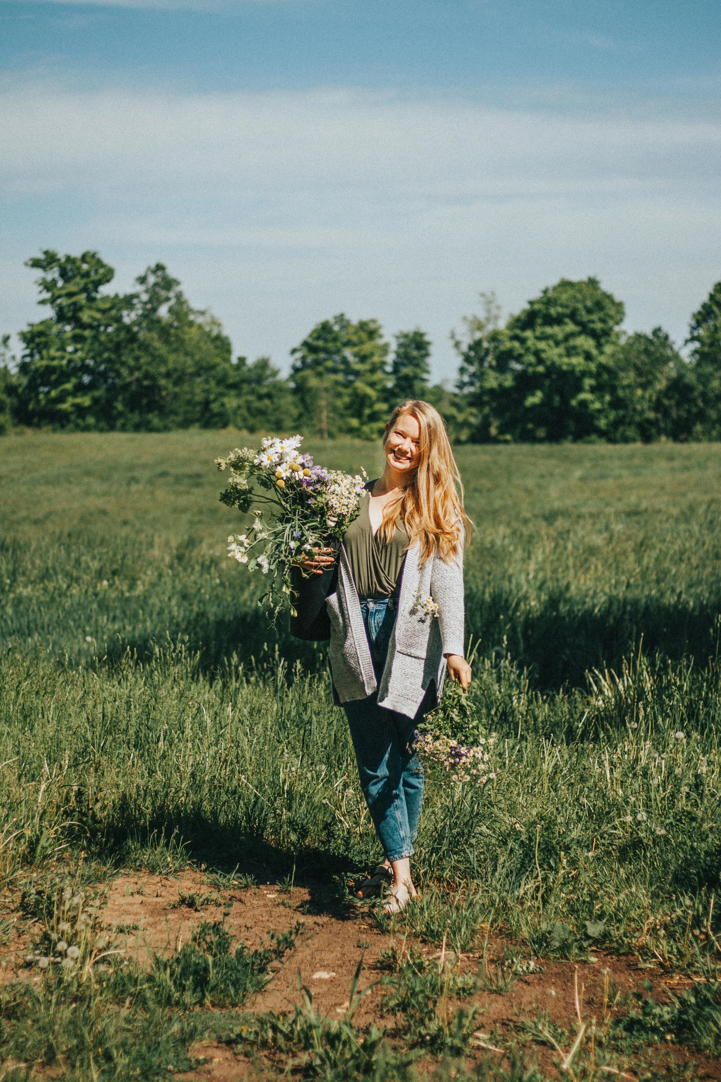 A woman with long blonde hair standing in a grassy field holding a large bouquet of wildflowers and a smaller bouquet in her hand, smiling.