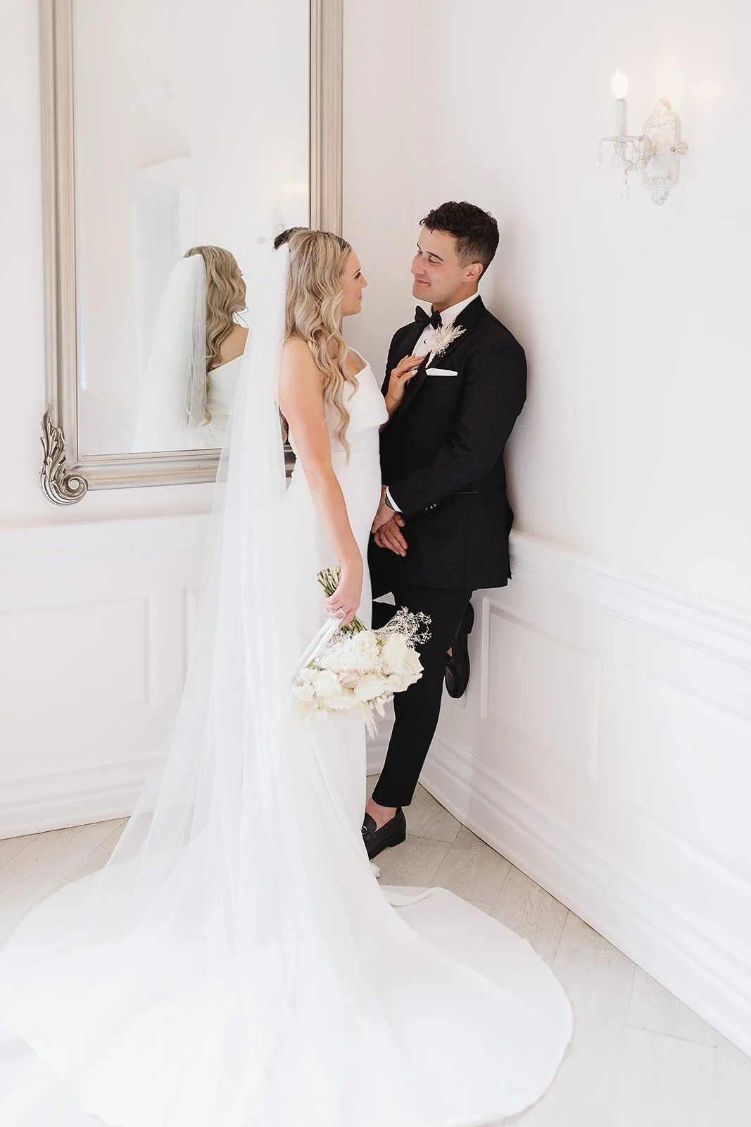 A bride and groom in formal wedding attire standing close together against a white wall with a large mirror and a wall sconce, looking into each other's eyes.