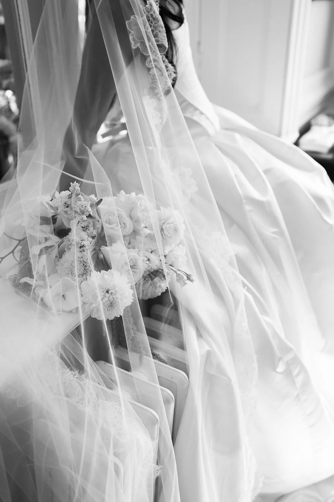 A bride sitting on a chair, holding a bouquet of flowers, wearing a wedding dress and veil, in black and white.