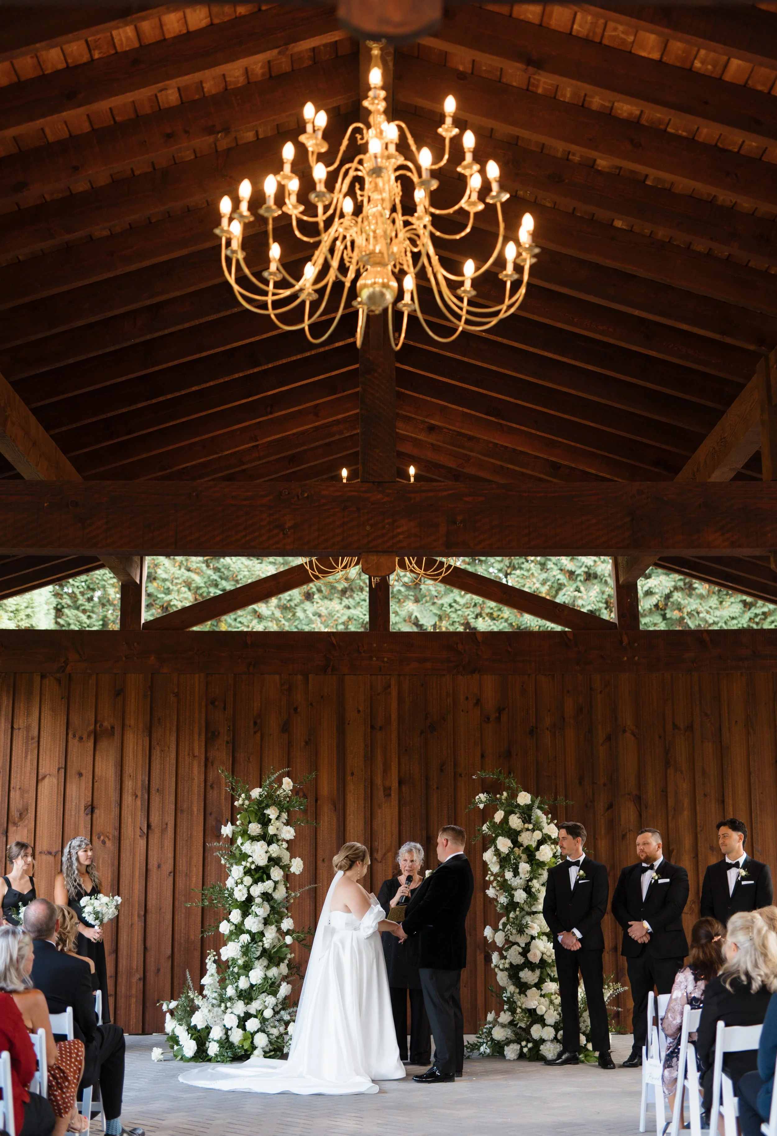 Wedding ceremony with bride and groom exchanging vows inside a wooden pavilion, surrounded by floral arrangements and guests watching.