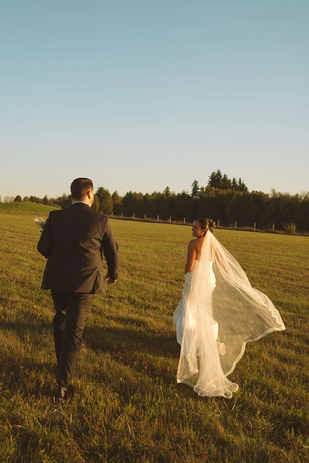 A bride and groom walk across a grassy field during sunset, with the bride smiling back at the groom.