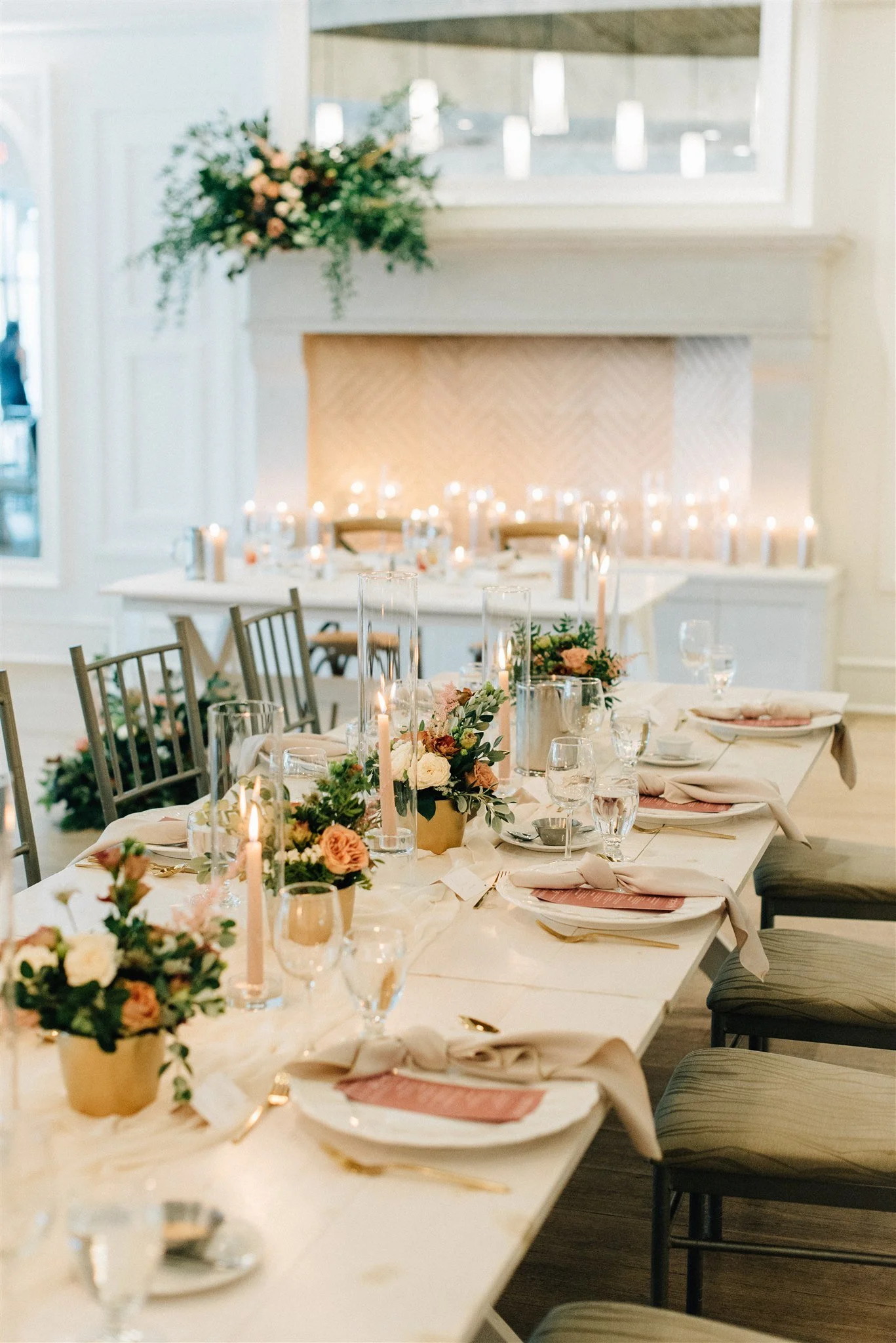 Elegant dining table set with floral centerpieces, pink napkins, candles, and glassware in a decorated room with a mantel and flowers.