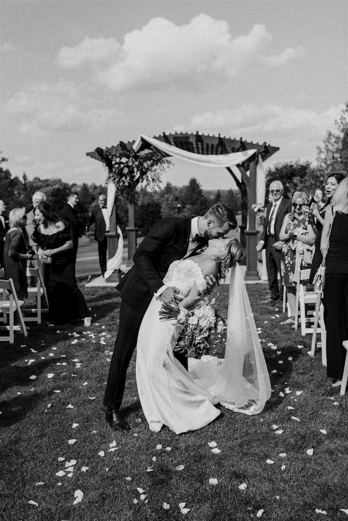 A newlywed couple sharing a kiss under a wedding arch, surrounded by guests at an outdoor wedding ceremony, in black and white.