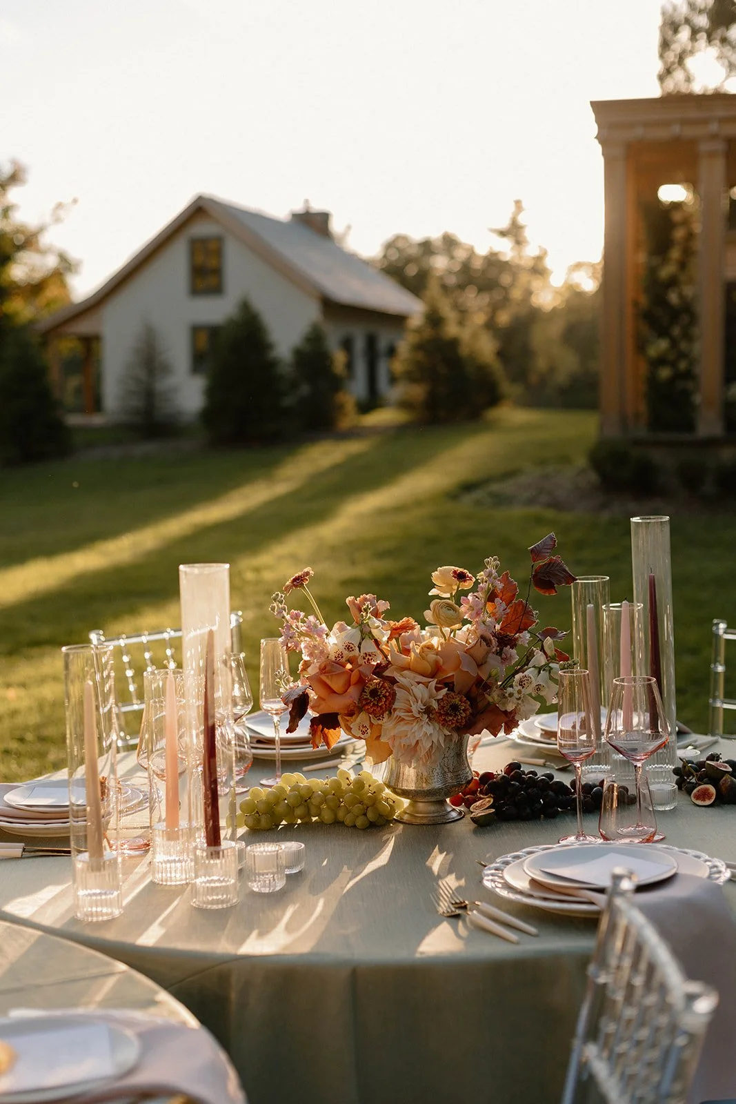 Outdoor event table set with plates, glasses, and a floral centerpiece, illuminated by warm sunlight.