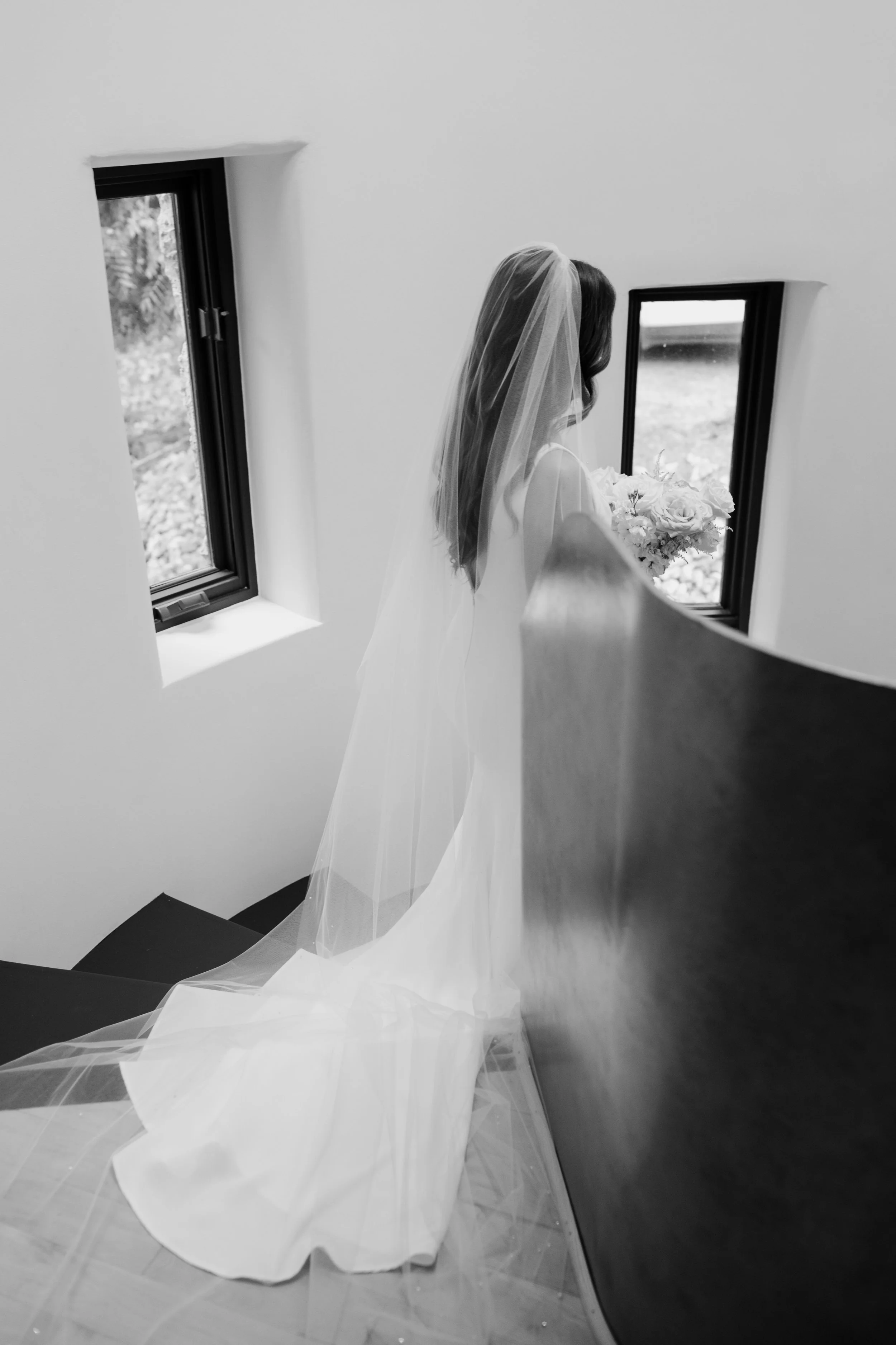 Black and white photo of a bride with a veil holding a bouquet, standing on a staircase near two small windows.