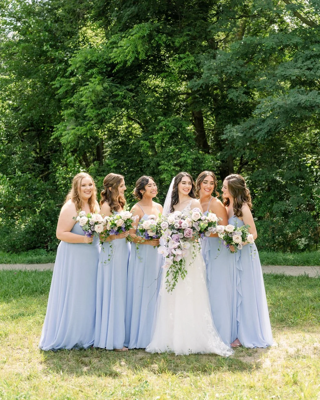Bridal party of six women standing outdoors in a green park, wearing light blue dresses, with the bride in a white wedding gown, holding bouquets of pink and white roses, smiling and talking to each other.