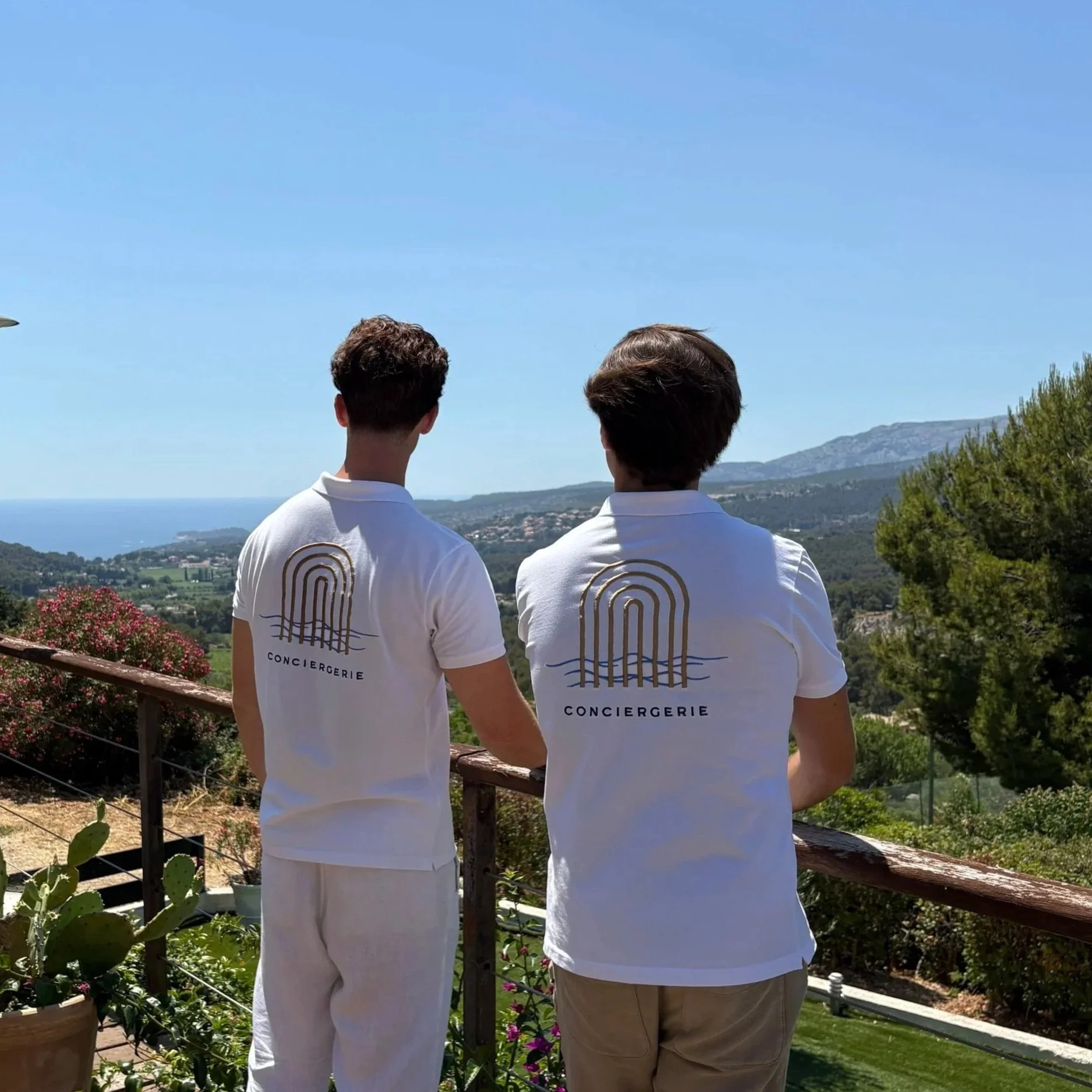 Deux jeunes hommes portant des t-shirts blancs avec un logo et le mot 'Conciergerie' regardent une vue panoramique sur la campagne, la mer et des montagnes, depuis une terrasse ornée de plantes.