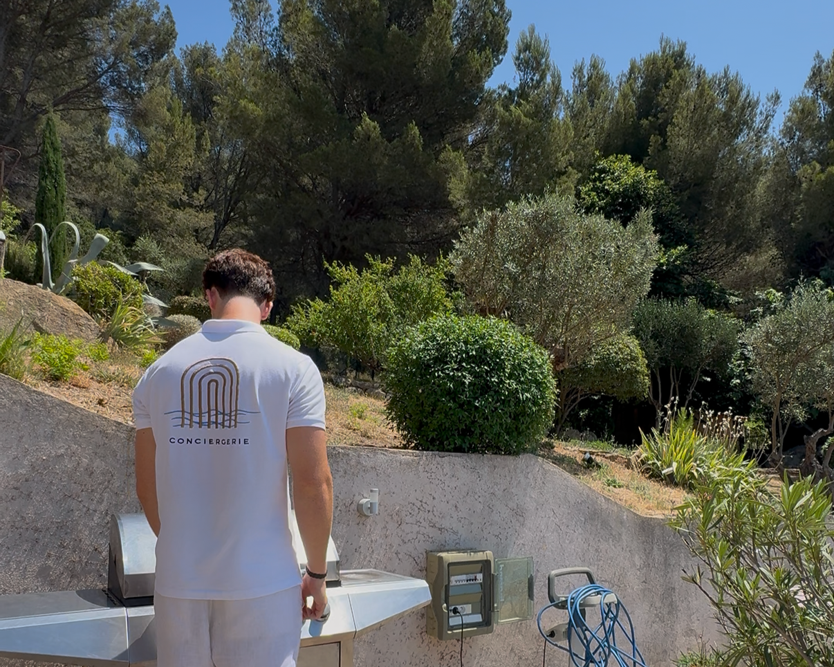 Homme vu de dos en uniforme blanc avec le logo 'Conciergerie' en train de préparer quelque chose sur un comptoir extérieur, avec un jardin en arrière-plan constitué d'arbustes et d'arbres, sous un ciel bleu.