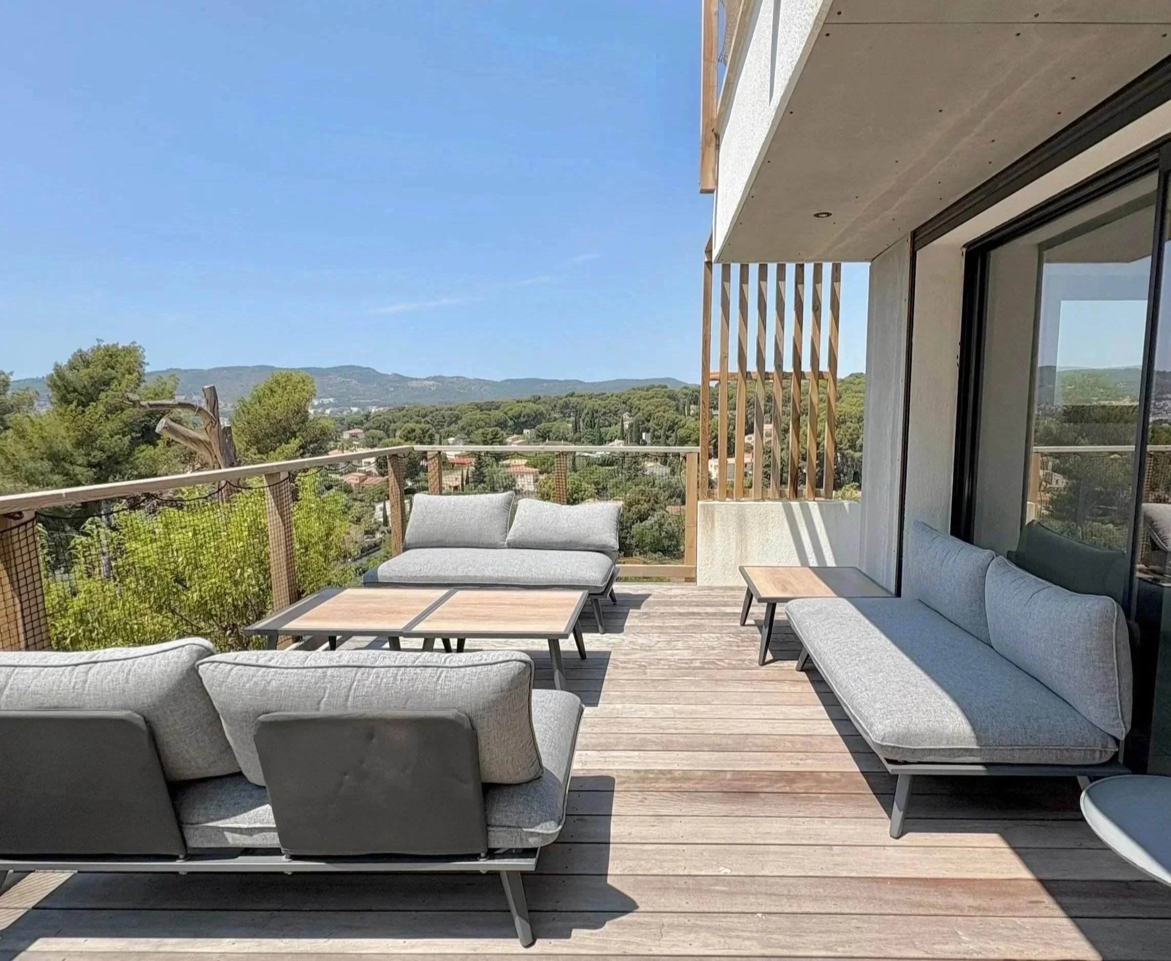 Balcon avec canapé et tables sous un ciel bleu, vue sur la végétation et les montagnes en arrière-plan.
