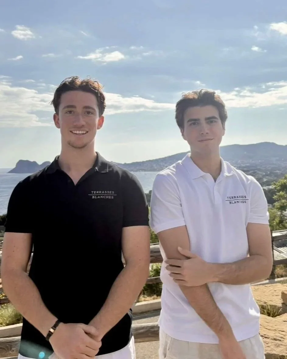 Deux jeunes hommes posant en extérieur avec vue sur la mer et des montagnes en arrière-plan, portant des polos avec l'inscription 'TERRASSES BLANCHES'.