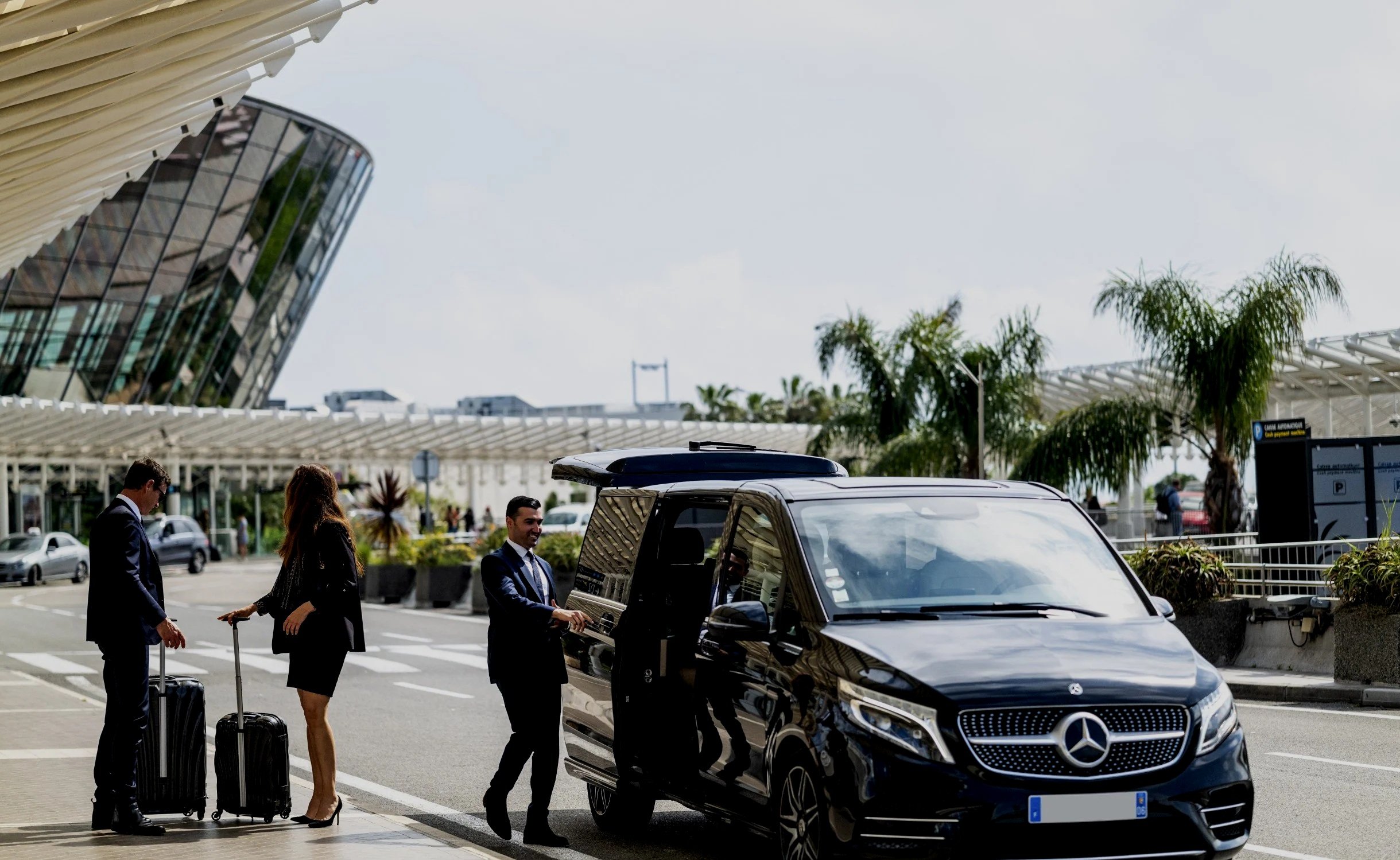 Trois personnes d'affaires se font déposer devant un terminal d'aéroport. Deux hommes en costume noir avec des valises, une femme en jupe et blazer noire qui monte dans une voiture noire Mercedes-Benz avec des employés en costume qui l'aident.