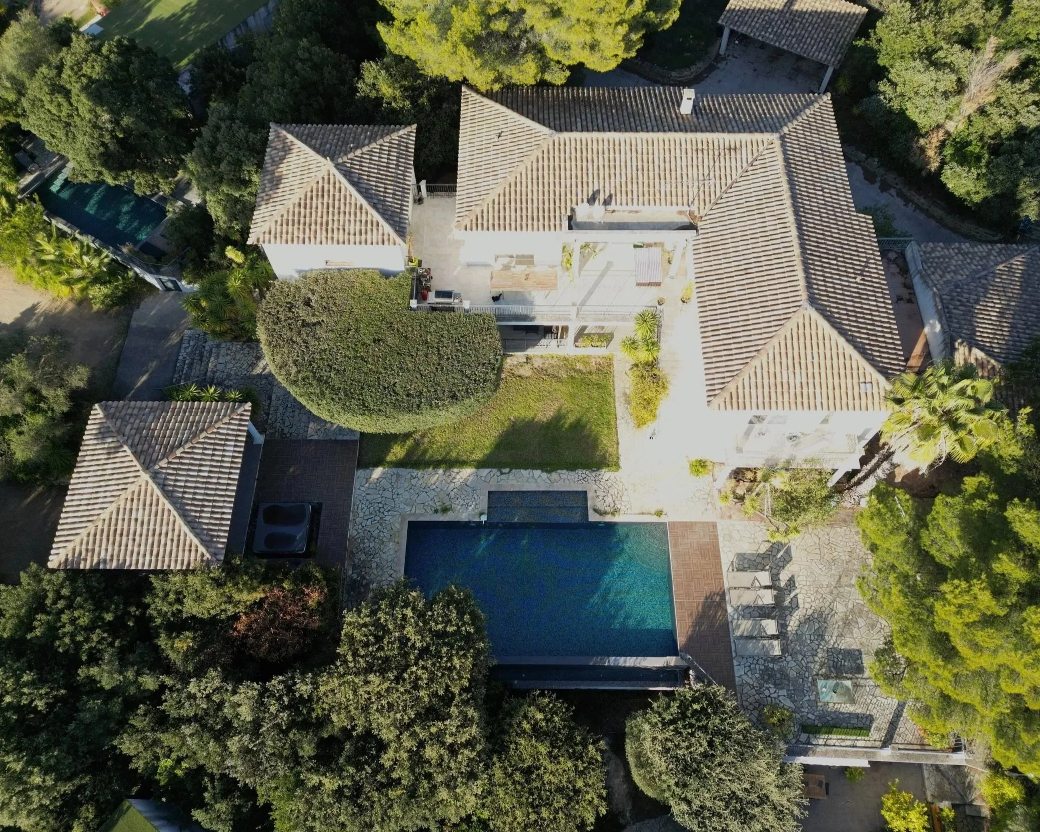 Vue aérienne d'une maison avec piscine, entourée d'arbres et de jardins, avec plusieurs toits en tuiles.