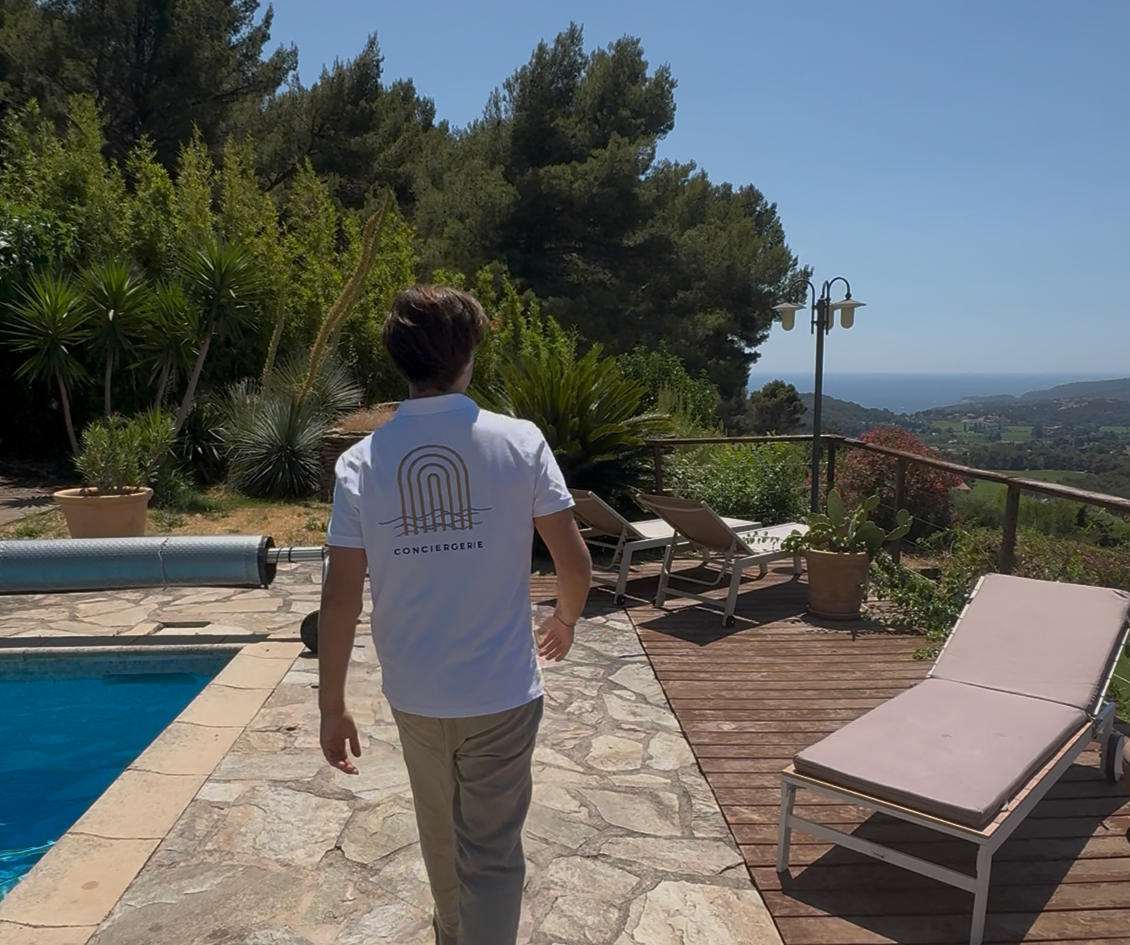Person avec t-shirt blanc portant un logo et le mot 'CONCIERGERIE', promenade autour d'une piscine avec mobilier de jardin, en terrasse avec vue sur la campagne et la mer par temps ensoleillé.