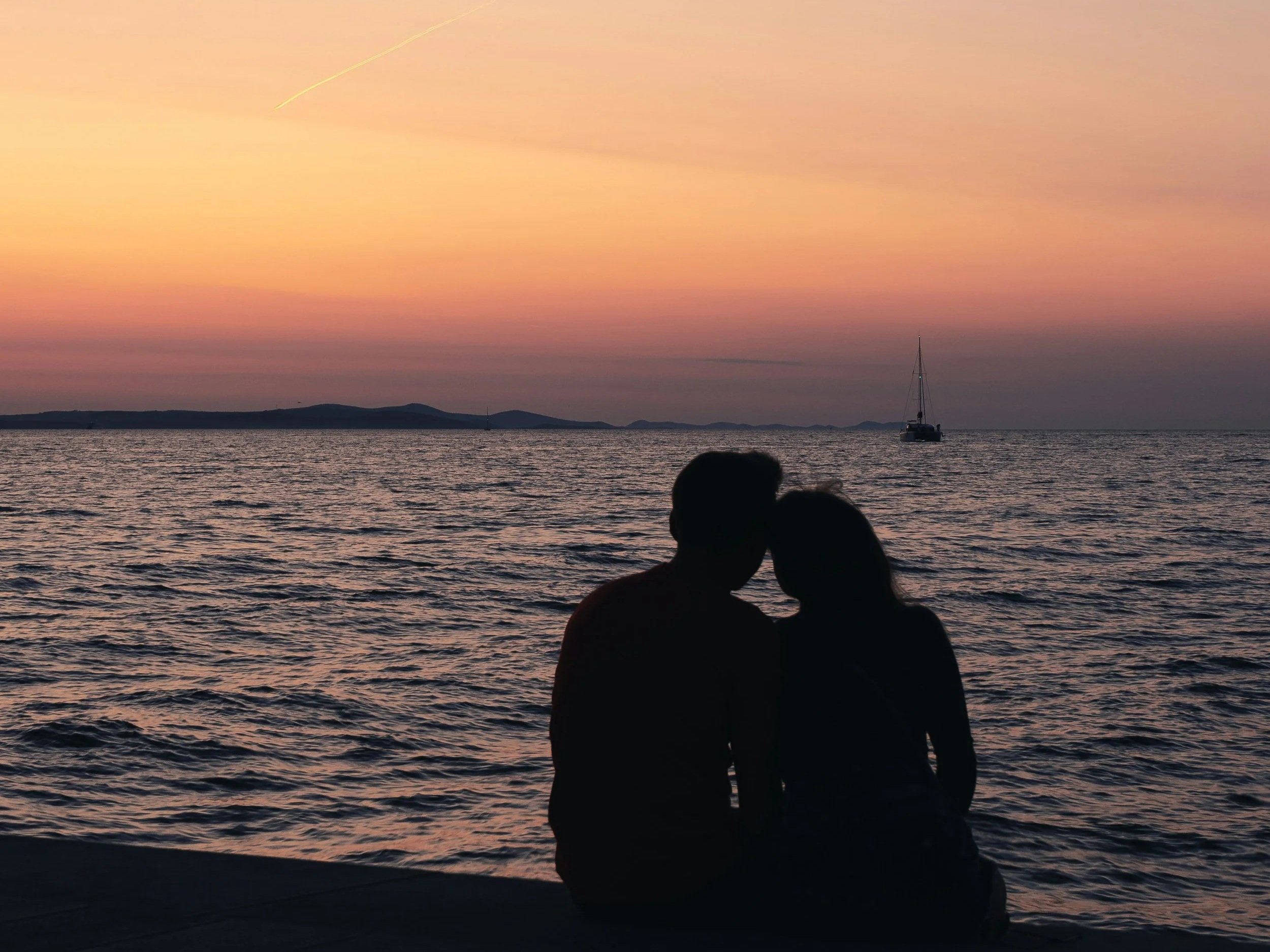 Silhouette d'un couple assis sur la plage au coucher du soleil, devant la mer, avec un bateau en arrière-plan et un ciel aux teintes roses et oranges.
