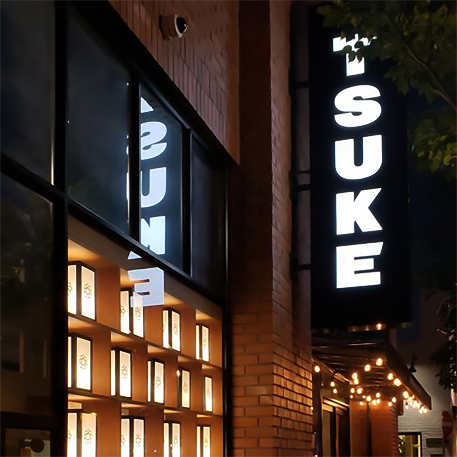 Exterior vertical sign that reads "TSUKE" during evening light, lanterns lit up at window
